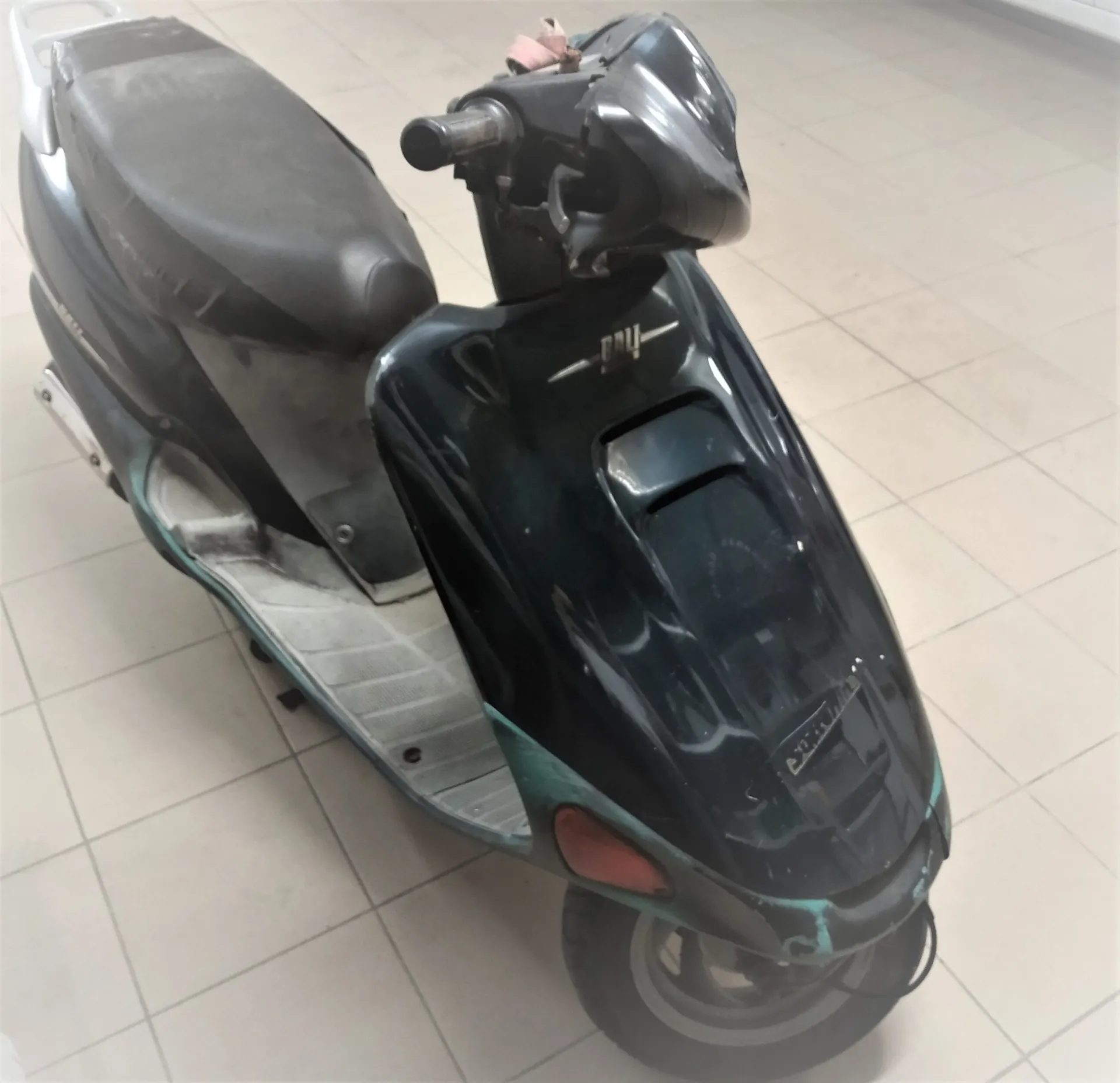 Czarny skuter zaparkowany w pomieszczeniu z jasną, kafelkową podłogą. Pojazd jest widoczny z perspektywy przedniej i bocznej, posiada czarne siedzenie oraz widoczne ślady zużycia i zarysowania na przedniej owiewce.