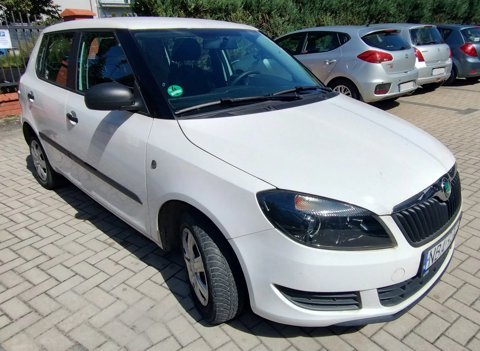 Biały samochód marki Skoda Fabia zaparkowany na kostce brukowej. Auto jest widoczne z przodu i z boku. W tle znajdują się inne zaparkowane samochody w kolorach srebrnym i niebieskim oraz zielone krzewy.