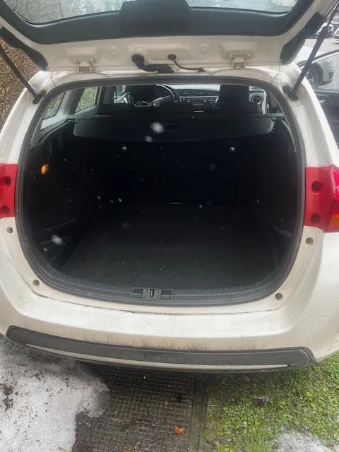 Otwarty bagażnik białego samochodu osobowego. Wnętrze bagażnika jest puste, widoczne są czarne tapicerowane ścianki i podłoga. W tle przez otwarte drzwi widoczna jest część wnętrza pojazdu, w tym kierownica i siedzenia. Samochód stoi na nawierzchni z resztkami śniegu i trawą. Na zewnątrz, po prawej stronie, częściowo widoczny jest inny pojazd. Bagażnik jest otwarty, a pokrywa uniesiona.