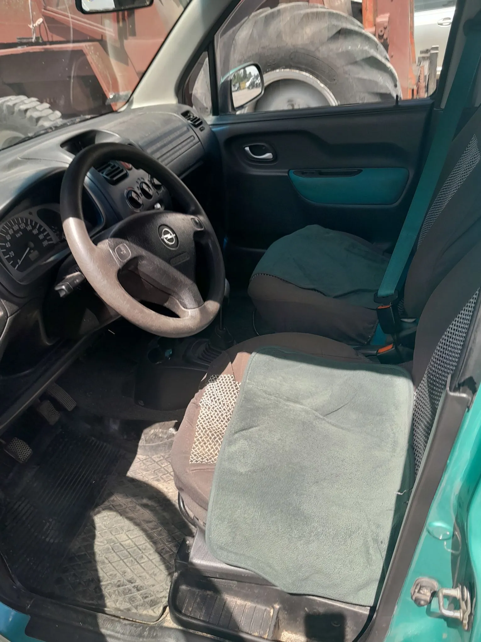 Wnętrze samochodu marki Opel, widok z perspektywy otwartych drzwi pasażera. Widoczna jest kierownica z logo producenta, deska rozdzielcza z zegarami oraz dwa przednie fotele z szarymi nakładkami ochronnymi na siedziskach. Podłoga wyłożona jest gumowymi dywanikami. Przez okno widać fragment dużej opony maszyny rolniczej.