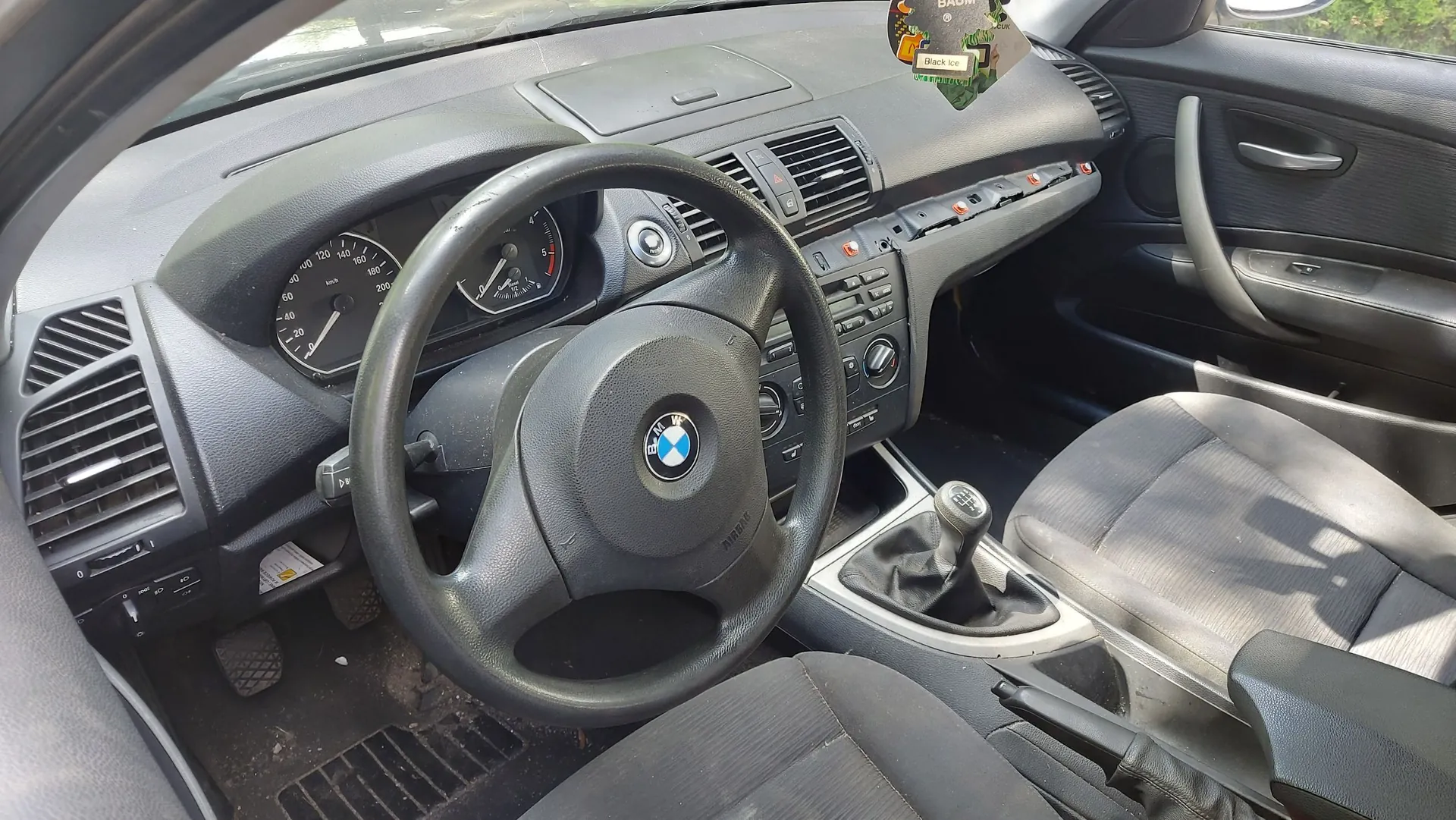 Wnętrze samochodu marki BMW z perspektywy kierowcy. Widoczna jest czarna kierownica z logo BMW w centrum, deska rozdzielcza z zegarami, nawiewami oraz konsolą środkową z drążkiem zmiany biegów. Pasażerska strona deski rozdzielczej jest częściowo rozebrana, odsłaniając wewnętrzne elementy konstrukcyjne. Widoczne są ciemne, materiałowe fotele oraz czarna tapicerka drzwi.