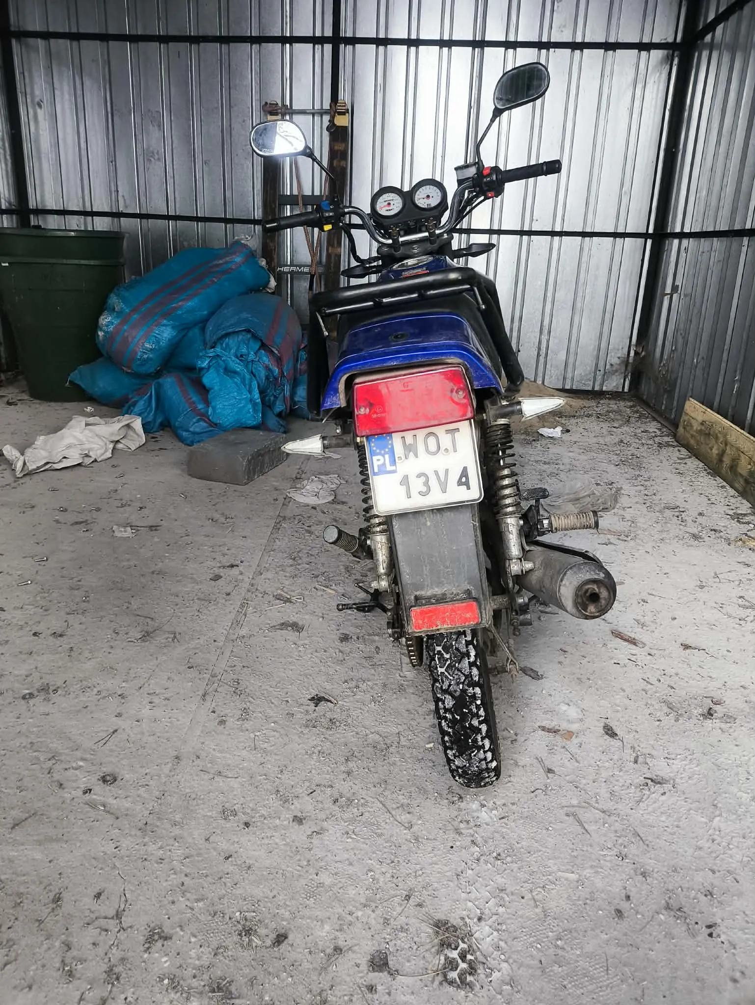Zdjęcie przedstawia niebieski motocykl widziany od tyłu, stojący w pomieszczeniu z metalowymi ścianami. Na motocyklu widoczna jest biała tablica rejestracyjna z napisem PL WOT 13V4, czerwone światła tylnego oraz dwa lusterka boczne. Po lewej stronie znajdują się zielony pojemnik i niebieskie worki z czerwonymi paskami, a na betonowej podłodze rozrzucone są różne przedmioty i śmieci. W tle metalowe ściany z pionowymi listwami.