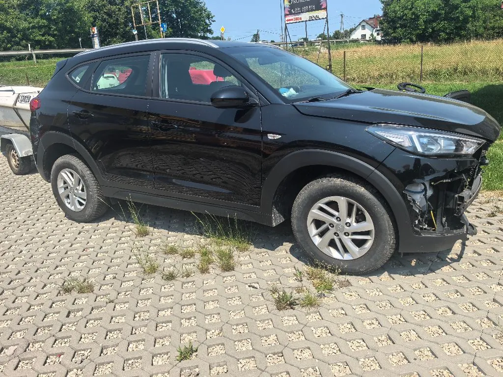 Czarny samochód typu SUV zaparkowany na kostce brukowej z dziurami, przez które wyrasta trawa. Pojazd jest uszkodzony w przedniej części po prawej stronie, gdzie brakuje zderzaka i widoczne są elementy konstrukcyjne. W tle widać zielone drzewa, krzewy oraz fragmenty budynków i tablic informacyjnych.