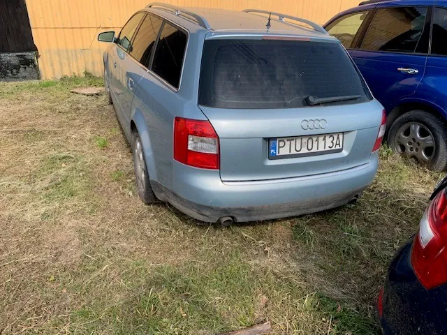 Jasnoniebieski samochód marki Audi w nadwoziu kombi, zaparkowany na trawiastym podłożu. Widoczny jest tył pojazdu z tablicą rejestracyjną o numerze PTU 0113A. Obok stoi fragment ciemnoniebieskiego samochodu, a w tle znajduje się żółta ściana budynku.