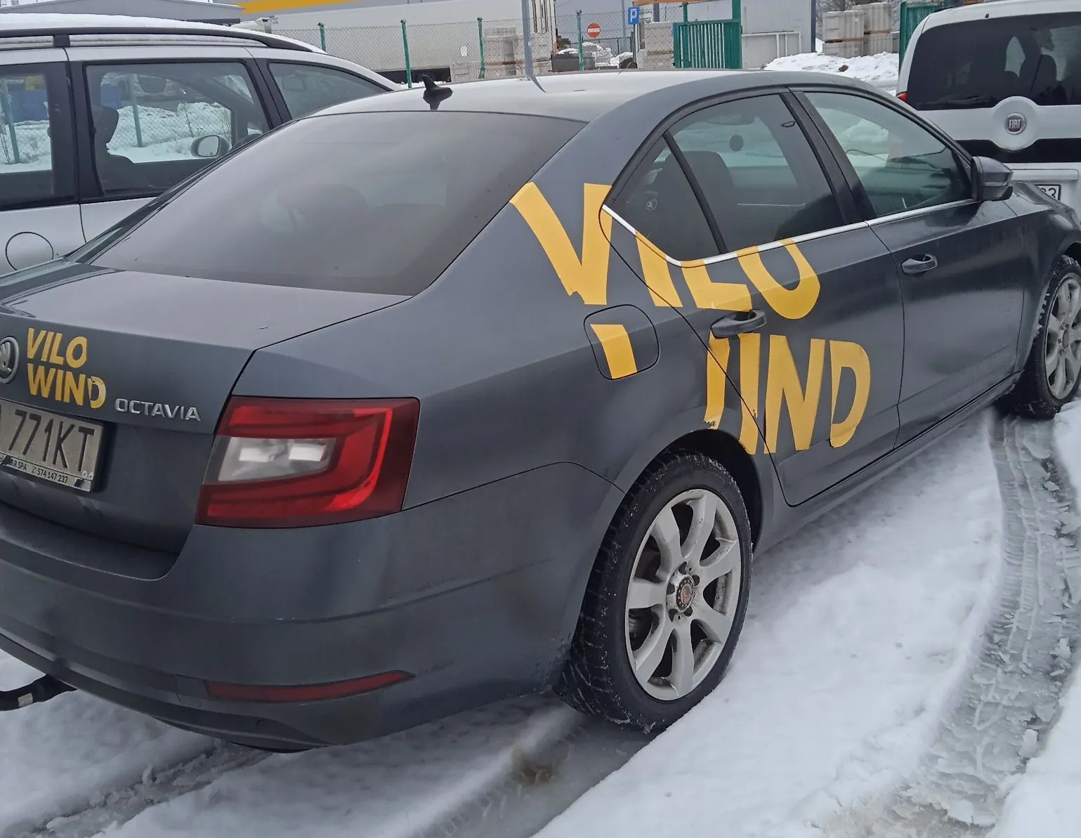 Szary samochód osobowy marki Skoda Octavia zaparkowany na zaśnieżonej nawierzchni. Na tylnym błotniku i drzwiach bocznych znajduje się duży, żółty napis "VILO WIND". Na klapie bagażnika widnieje mniejszy żółty napis "VILO WIND" oraz napis "OCTAVIA". Tablica rejestracyjna zawiera tekst "771 KT".