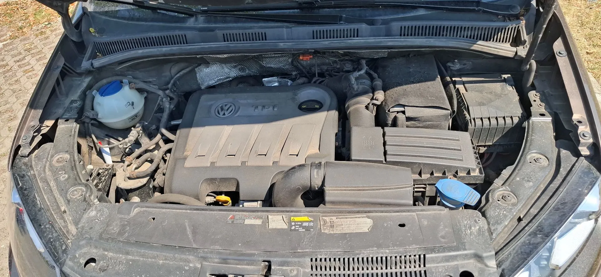 Widok z góry na komorę silnika samochodu marki Volkswagen. W centrum znajduje się plastikowa osłona silnika z logo VW i oznaczeniem TPI. Po lewej stronie widoczny jest biały zbiornik z niebieską zakrętką, po prawej czarne skrzynki z elektroniką oraz niebieska zakrętka zbiornika spryskiwaczy. Całość jest zakurzona.