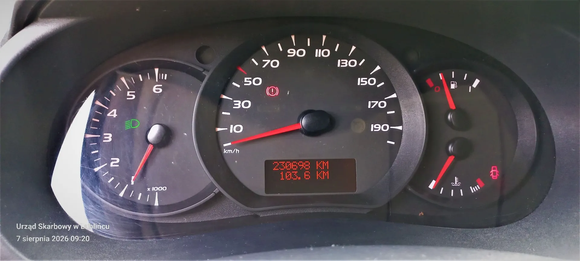 Zdjęcie deski rozdzielczej samochodu. W centrum znajduje się prędkościomierz z zaznaczonymi wartościami od 10 do 190 km/h, z czerwonymi wskazówkami. Poniżej prędkościomierza znajduje się cyfrowy wyświetlacz z czerwonym tekstem: '238698 KM' oraz '103.6 KM'. Po lewej stronie widoczny jest obrotomierz z zaznaczonymi wartościami od 1 do 6 (x 1000) i zapaloną zieloną kontrolką świateł. Po prawej stronie znajdują się wskaźniki poziomu paliwa oraz temperatury silnika. W lewym dolnym rogu obrazu znajduje się biały napis: 'Urząd Skarbowy w Lublińcu 7 sierpnia 2026 09:20'.