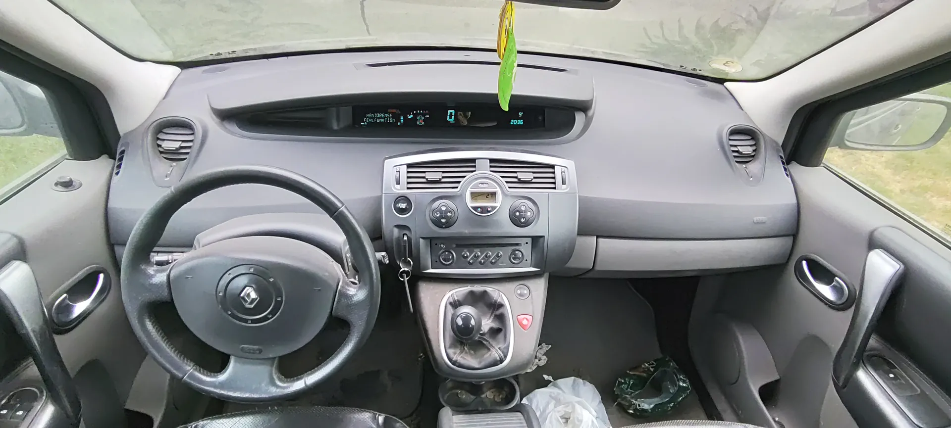 Wnętrze samochodu z widokiem na deski rozdzielczą, kierownicę z logo Renault, centralną konsolę z przyciskami i wyświetlaczem, dźwignią zmiany biegów oraz przedmiotami na podłodze (plastikowa torba, zielony przedmiot). Na wyświetlaczu deski rozdzielczej widoczne są cyfry 0 i 2036 oraz inne symbole. W lusterku wstecznym wiszą żółto-zielone ozdoby. Drzwi po obu stronach z uchwytami i oknami, przez które widać trawę na zewnątrz.