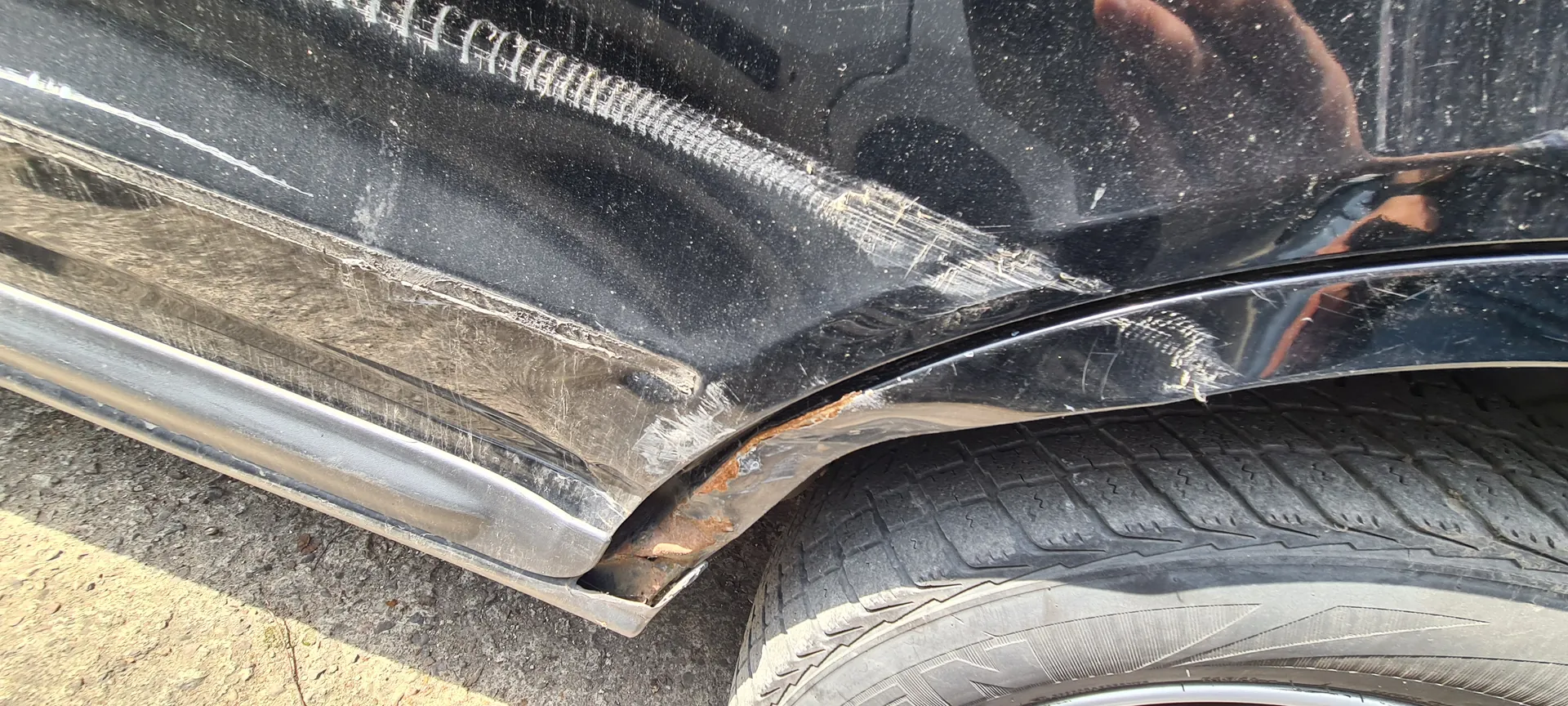 Czarny samochód z widocznymi uszkodzeniami na tylnym zderzaku, w tym zadrapaniami i rdzą w okolicy nadkola. Widoczna opona z wyraźnym wzorem bieżnika oraz asfaltowa powierzchnia z żółtą linią. W odbiciu na karoserii widoczna ręka trzymająca aparat.