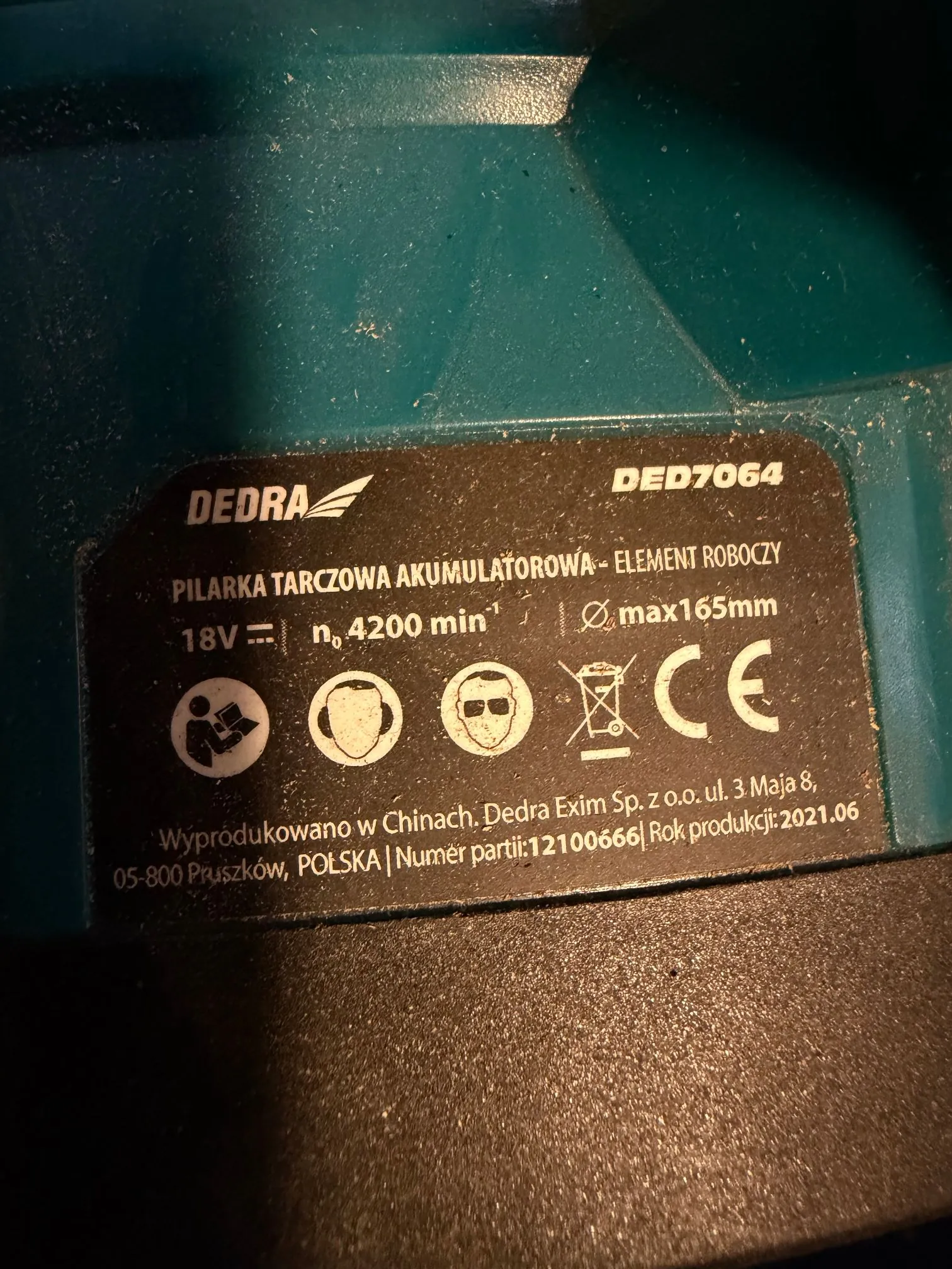 Zbliżenie na tabliczkę znamionową urządzenia marki DEDRA, model DED7064. Tekst na tabliczce brzmi: 'PILARKA TARCZOWA AKUMULATOROWA - ELEMENT ROBOCZY', '18V', 'n 4200 min-1', 'Ø max 165mm'. Widoczne są piktogramy dotyczące bezpieczeństwa: instrukcja obsługi, ochrona słuchu, ochrona oczu oraz symbol przekreślonego kosza na śmieci i znak CE. Na dole znajduje się napis: 'Wyprodukowano w Chinach. Dedra Exim Sp. z o.o. ul. 3 Maja 8, 05-800 Pruszków, POLSKA | Numer partii: 12100666 | Rok produkcji: 2021.06'. Tabliczka jest przyklejona do ciemnozielonej, plastikowej obudowy urządzenia.