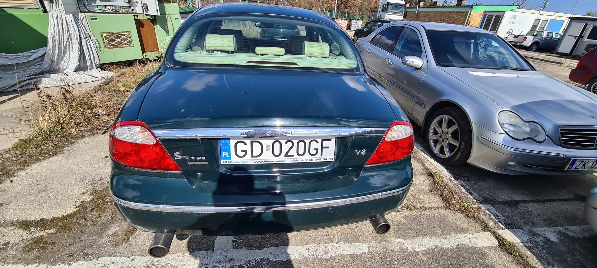 Z tyłu widoczny ciemnozielony samochód Jaguar S-Type V8 z tablicą rejestracyjną GD 020GF (PL), na której widnieje napis HLW-Serwis www.hlwservis.pl oraz numer telefonu. Obok zaparkowany srebrny samochód Mercedes-Benz z częściowo widoczną tablicą ZK 0. Tło: betonowy parking, zielona budowla, inne pojazdy i budynki. Na asfalcie widoczne białe linie parkingu.