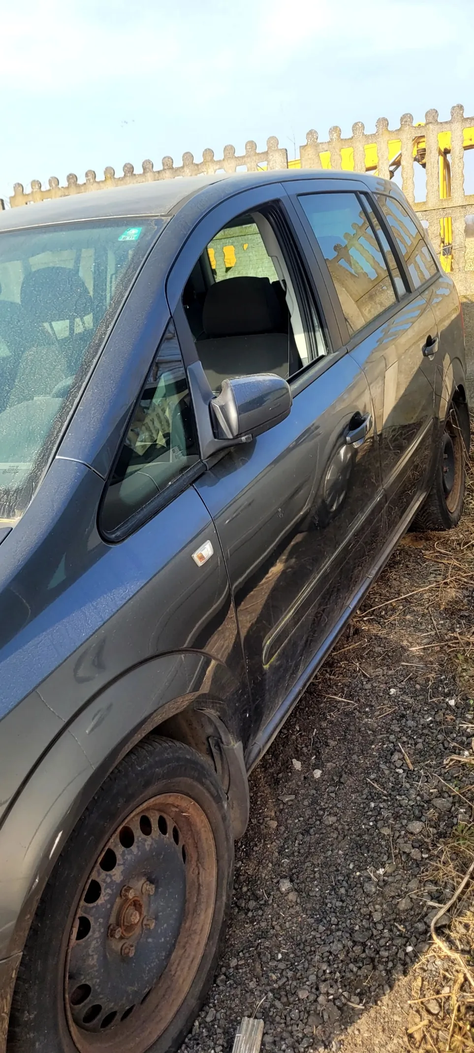 Ciemnoszary SUV zaparkowany na żwirowej powierzchni. Samochód ma zabrudzone koła z widoczną rdzą na felgach. W tle betonowy płot z żółtymi elementami konstrukcyjnymi oraz jasne niebo. Wnętrze pojazdu z widocznymi siedzeniami i przednią szybą z kroplami wody.