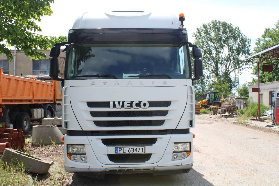 Widok z przodu białej ciężarówki marki Iveco Stralis zaparkowanej na piaszczystym placu. Pojazd posiada tablicę rejestracyjną o numerze PL 63471. Po lewej stronie widoczna jest pomarańczowa naczepa wywrotki, a w tle po prawej stronie żółta koparka oraz budynki gospodarcze. Wokół znajdują się drzewa i elementy betonowe.