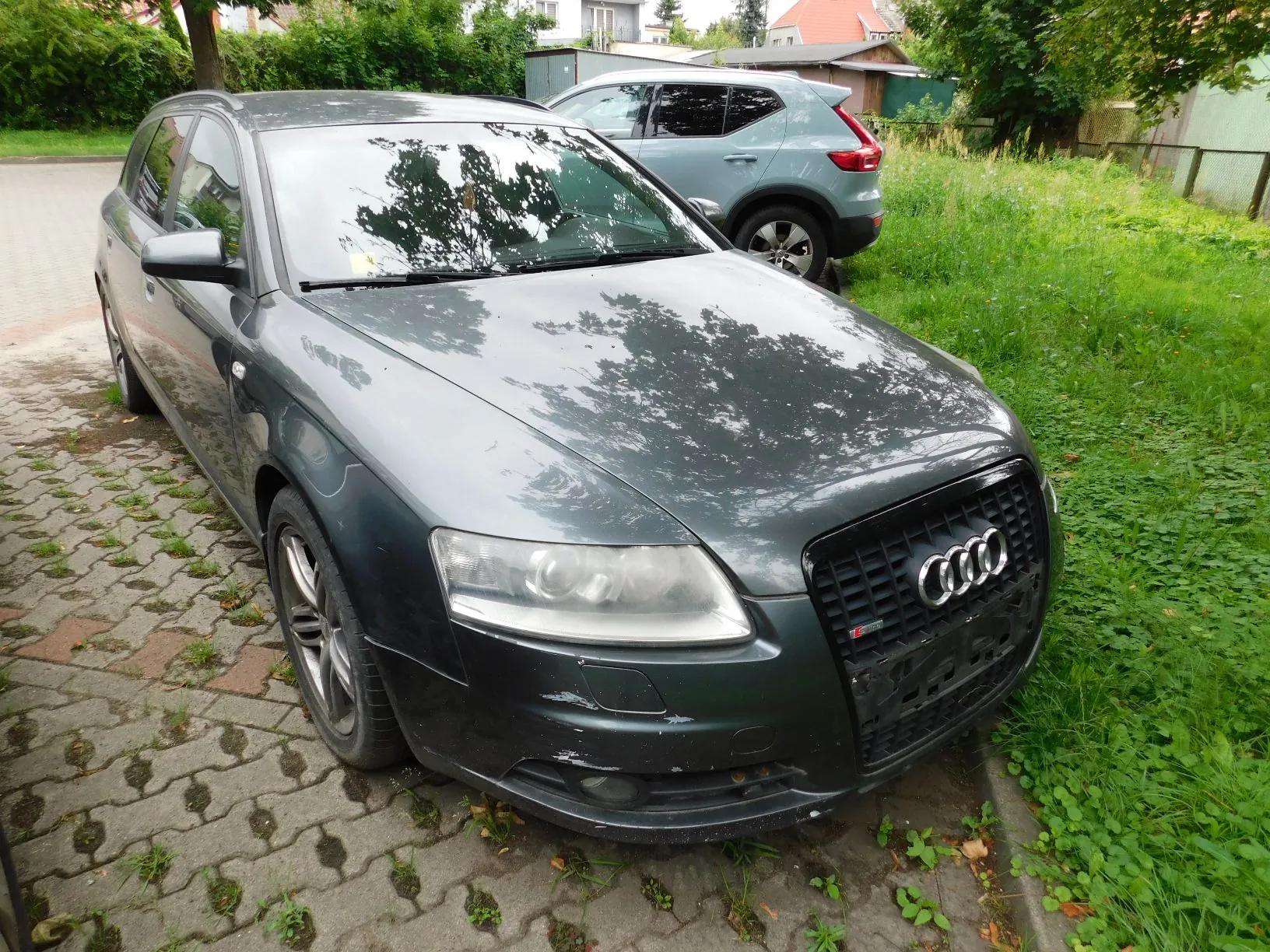 Szary samochód marki Audi zaparkowany na kostce brukowej obok trawnika. Auto ma widoczne uszkodzenia na przednim zderzaku po prawej stronie. W tle widoczny jest drugi, srebrny samochód oraz zieleń i fragmenty budynków.