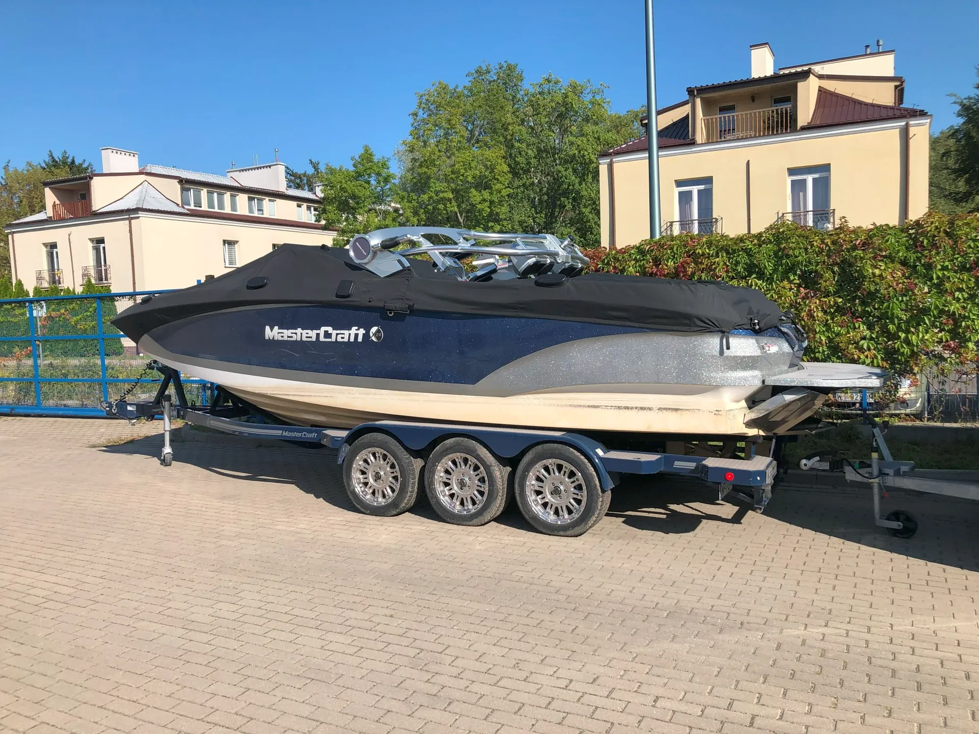 Motorówka marki MasterCraft na trójosiowej przyczepie, zaparkowana na kostce brukowej. Łódź jest przykryta czarnym pokrowcem, ma niebiesko-srebrny kadłub z białym dołem i widocznym napisem 'MasterCraft' na boku. W tle znajdują się dwa jasne domy jednorodzinne, zielone drzewa i krzewy pod bezchmurnym, niebieskim niebem.
