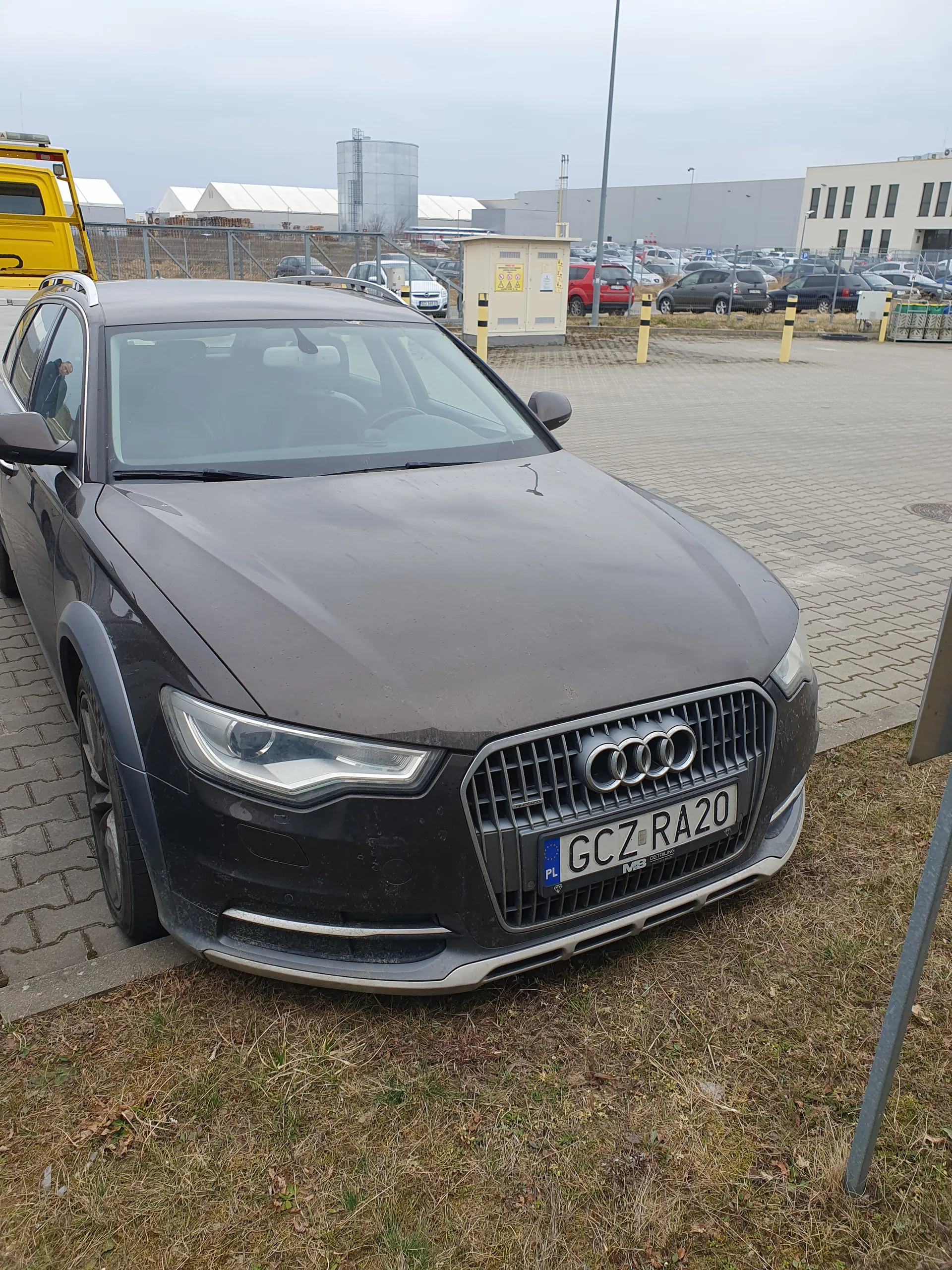Szary samochód Audi zaparkowany na brukowanej powierzchni obok trawy. Widoczna tablica rejestracyjna GCZ RA20 z niebieskim pasem UE. W tle budynki, inne zaparkowane samochody, żółta ciężarówka oraz jasne niebo.