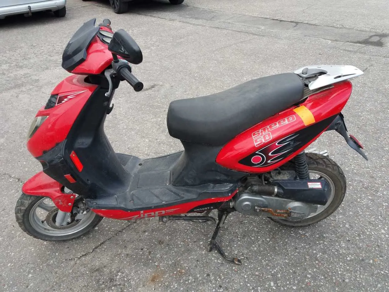 Czerwono-czarny skuter marki Zipp z napisem 'Speed 50' na boku, czarnym siedzeniem i srebrnym bagażnikiem tylnym. Skuter stoi na asfaltowej powierzchni, w tle częściowo widoczne są inne pojazdy.