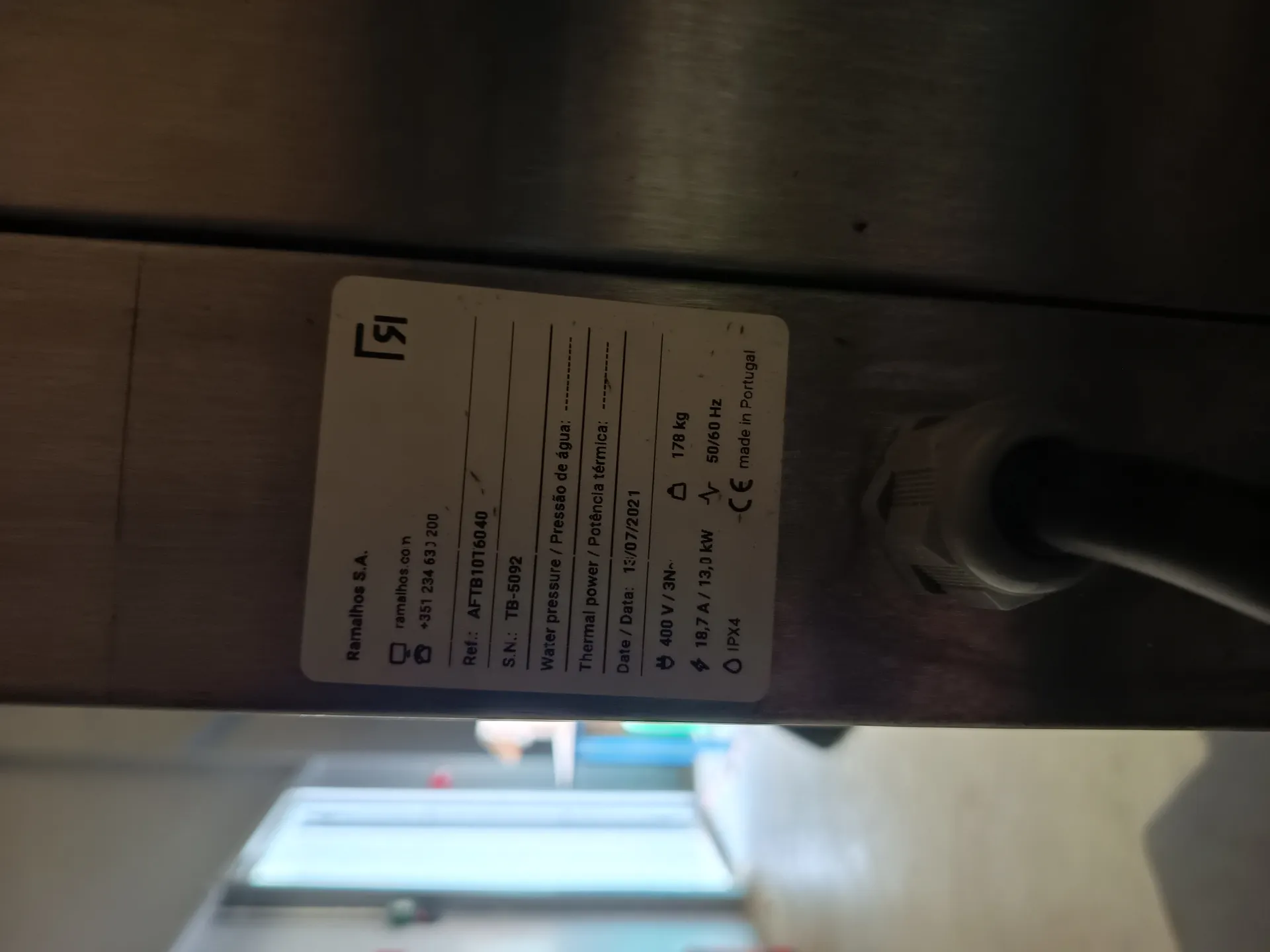 Biała etykieta z czarnym tekstem na metalowej powierzchni. Etykieta zawiera informacje o firmie Ramalhos S.A., adresie internetowym ramalhos.com, numerze telefonu +351 234 631 200, numerze referencyjnym AFTB1076040, numerze seryjnym TB-5092, parametrach takich jak ciśnienie wody / Pressão de água, moc termiczna / Potência térmica, data 13/07/2021, napięciu 400 V / 3N~, prądzie 18,7 A / 13,0 kW, częstotliwości 50/60 Hz, stopniu ochrony IPX4, masie 178 kg oraz oznaczeniu CE i 'made in Portugal'. W tle rozmyty widok pomieszczenia z drzwiami i przedmiotami. Poniżej etykiety widoczny jest czarny kabel podłączony do metalowej powierzchni.