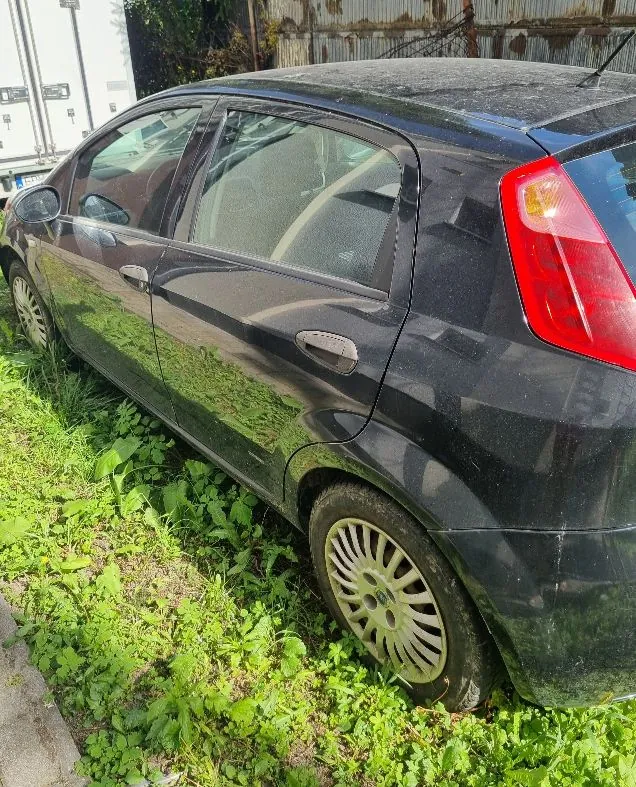 Zdjęcie przedstawia tylną, lewą część czarnego samochodu osobowego typu hatchback, zaparkowanego na trawiastym poboczu. Pojazd jest zakurzony, widoczna jest tylna lampa w kolorze czerwonym oraz koło z wieloramienną felgą. W tle widać fragment białej naczepy ciężarówki oraz metalowy płot.