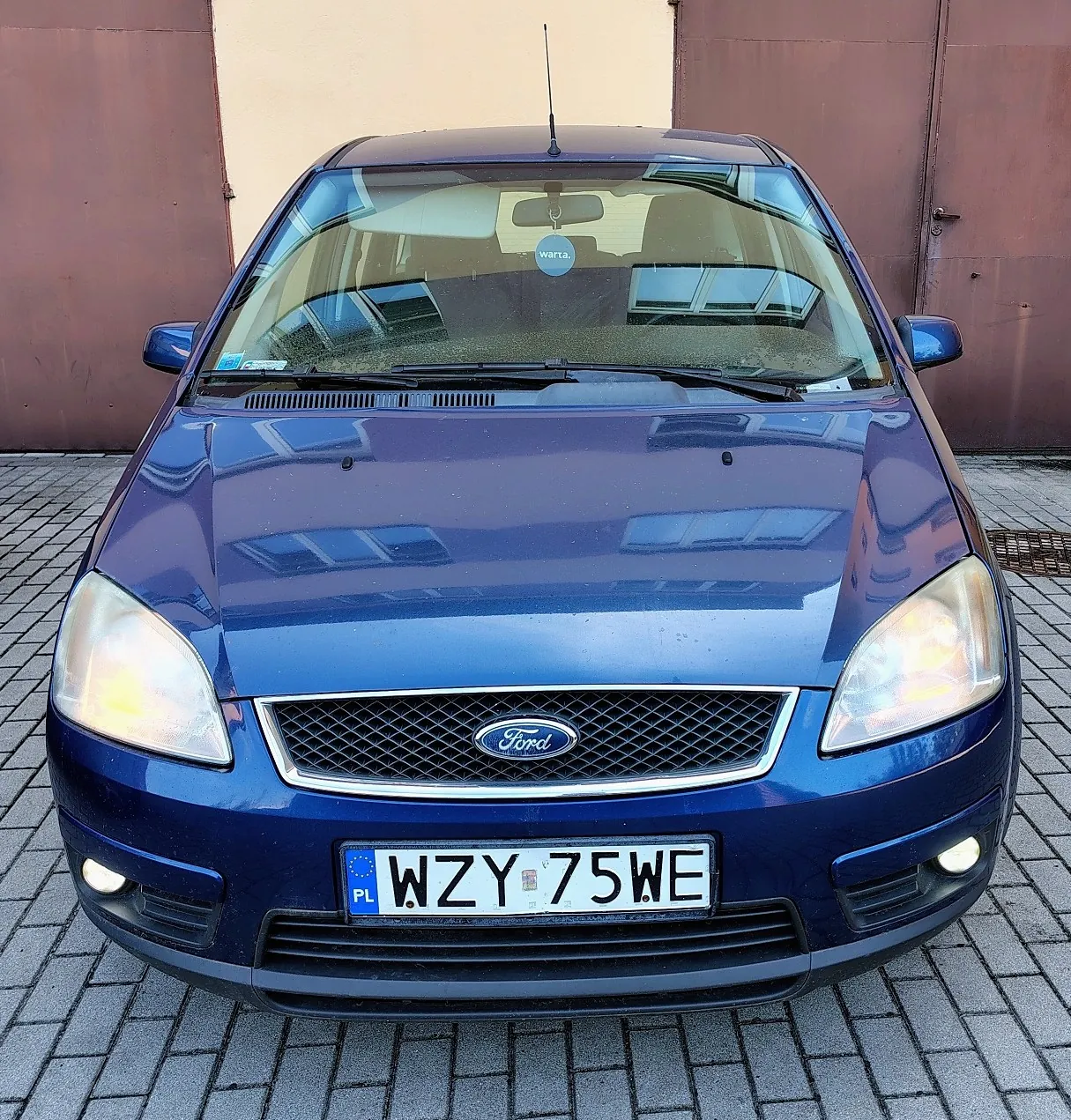 Niebieski samochód marki Ford zaparkowany na kostce brukowej przed brązową ścianą. Widoczny jest przód pojazdu z logo Ford na grillu oraz polska tablica rejestracyjna o numerze WZY 75WE.