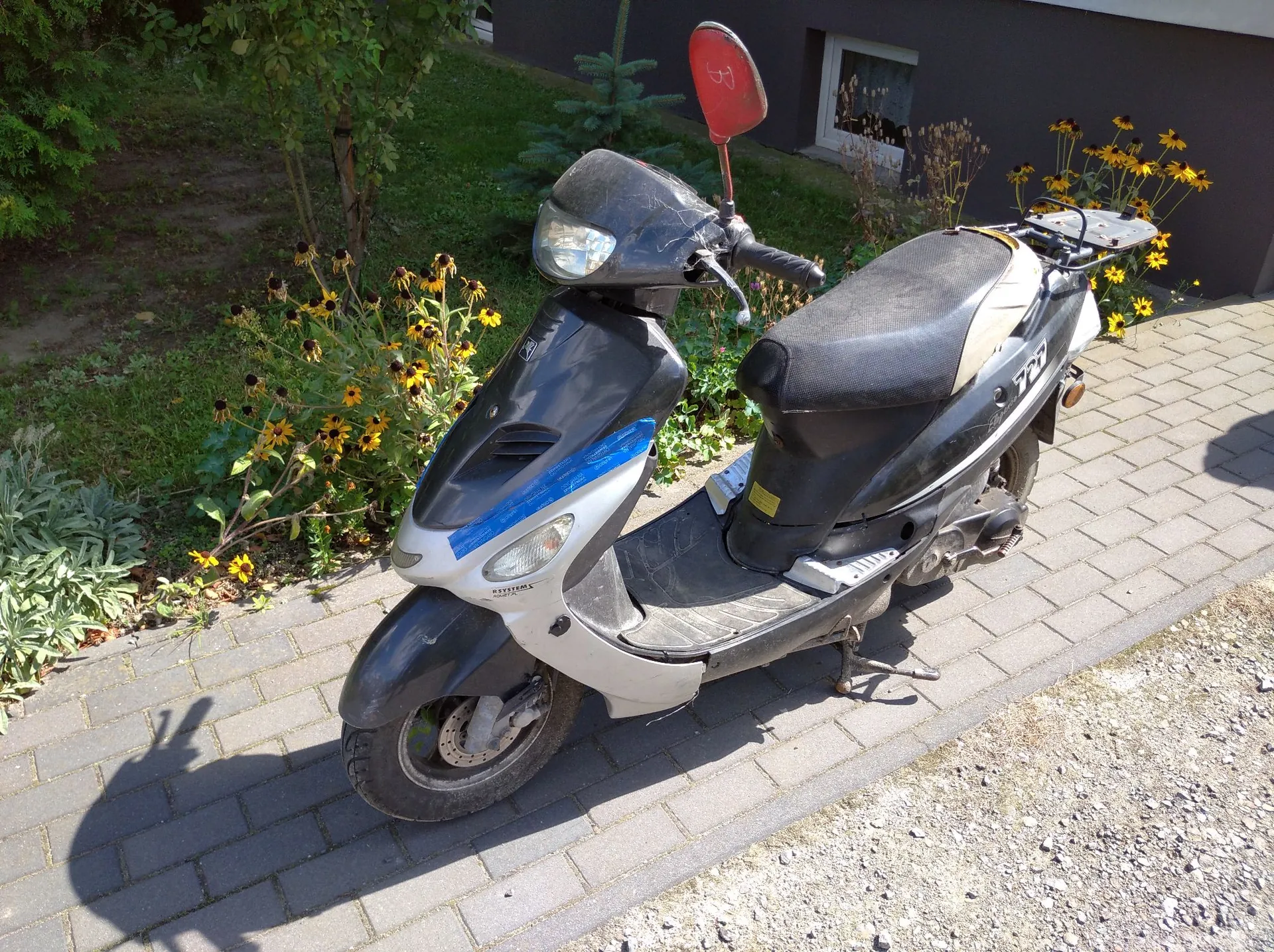 Czarny skuter marki Citroën zaparkowany na kostce brukowej. Pojazd ma srebrne i niebieskie akcenty na przedniej obudowie oraz czerwone lusterko wsteczne. W tle widoczne są zielone krzewy, żółte kwiaty oraz fragment szarego budynku z oknem.