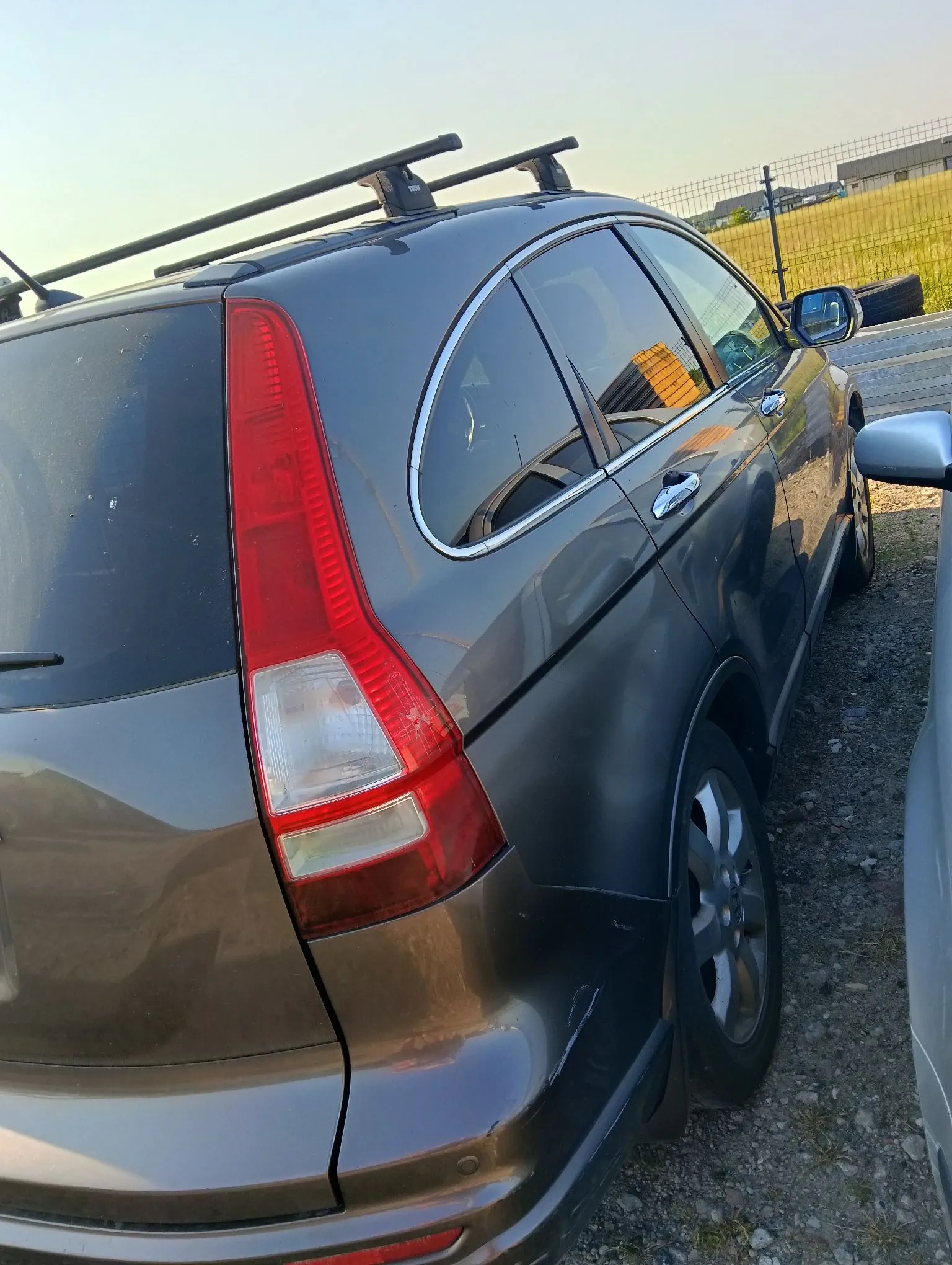 Zdjęcie tylnej części szarego samochodu typu kombi z boku. Pojazd posiada zamontowany na dachu czarny bagażnik dachowy. Widoczna jest pionowa tylna lampa w kolorach czerwonym i białym oraz srebrna obramowanie tylnej szyby. Samochód stoi na żwirowym podłożu obok innego, jasnego pojazdu.