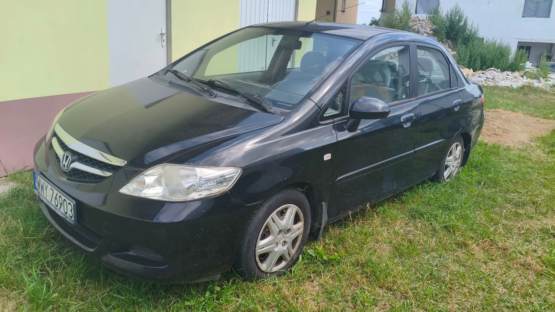 Czarny samochód osobowy marki Honda zaparkowany na trawie przed jasnym budynkiem. Widoczna jest przednia część i prawy bok auta. Samochód posiada tablicę rejestracyjną o numerze WNY 76903.