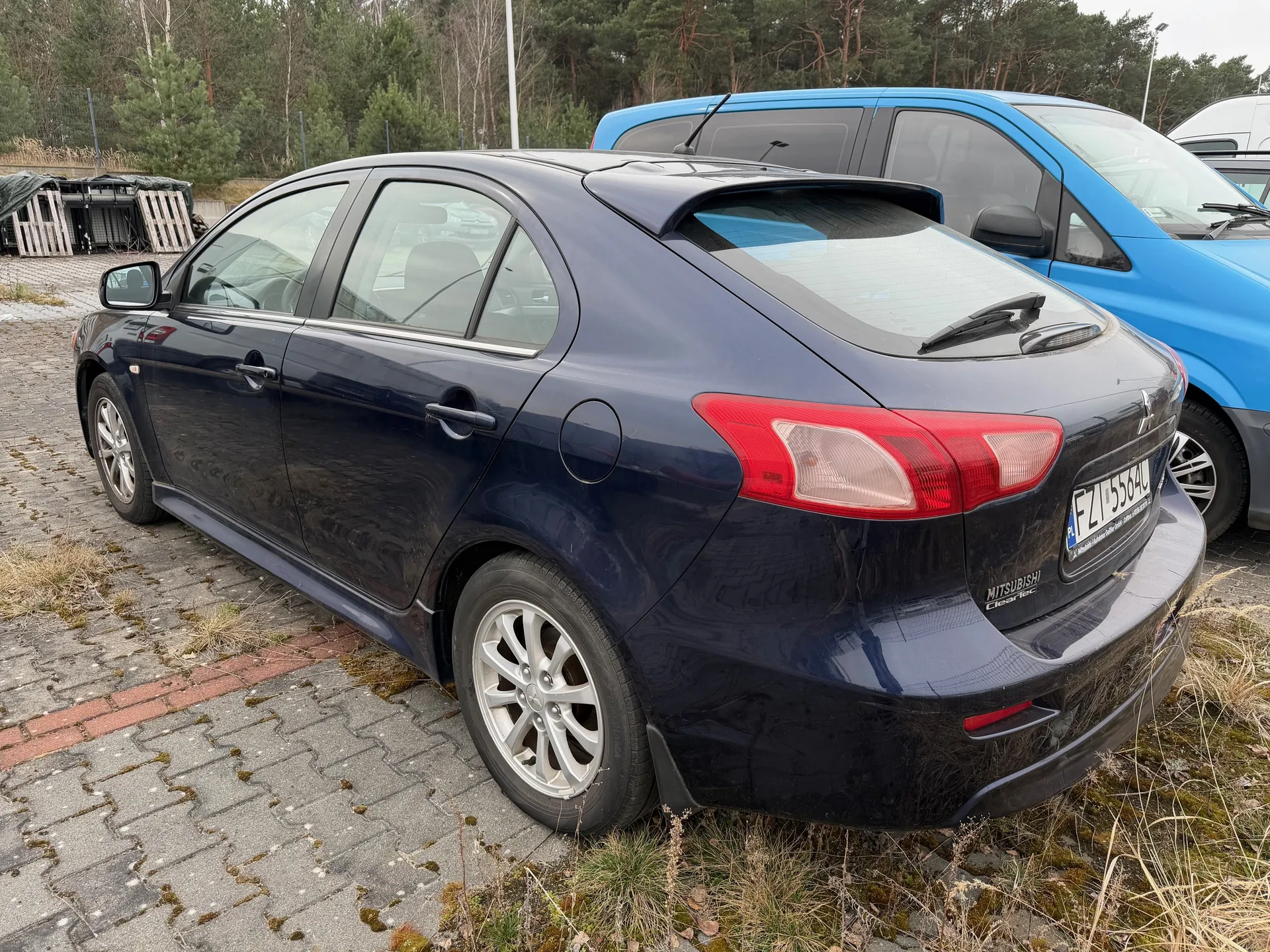 Ciemnoniebieski samochód osobowy Mitsubishi z tablicą rejestracyjną FZ 5564U zaparkowany na brukowanej powierzchni. Widoczne są czerwone światła tylne, srebrne felgi oraz napisy 'MITSUBISHI' i 'ClearTec' na bagażniku. W tle niebieski samochód dostawczy, drzewa, palety drewniane przykryte folią oraz sucha trawa na brzegu parkingu.