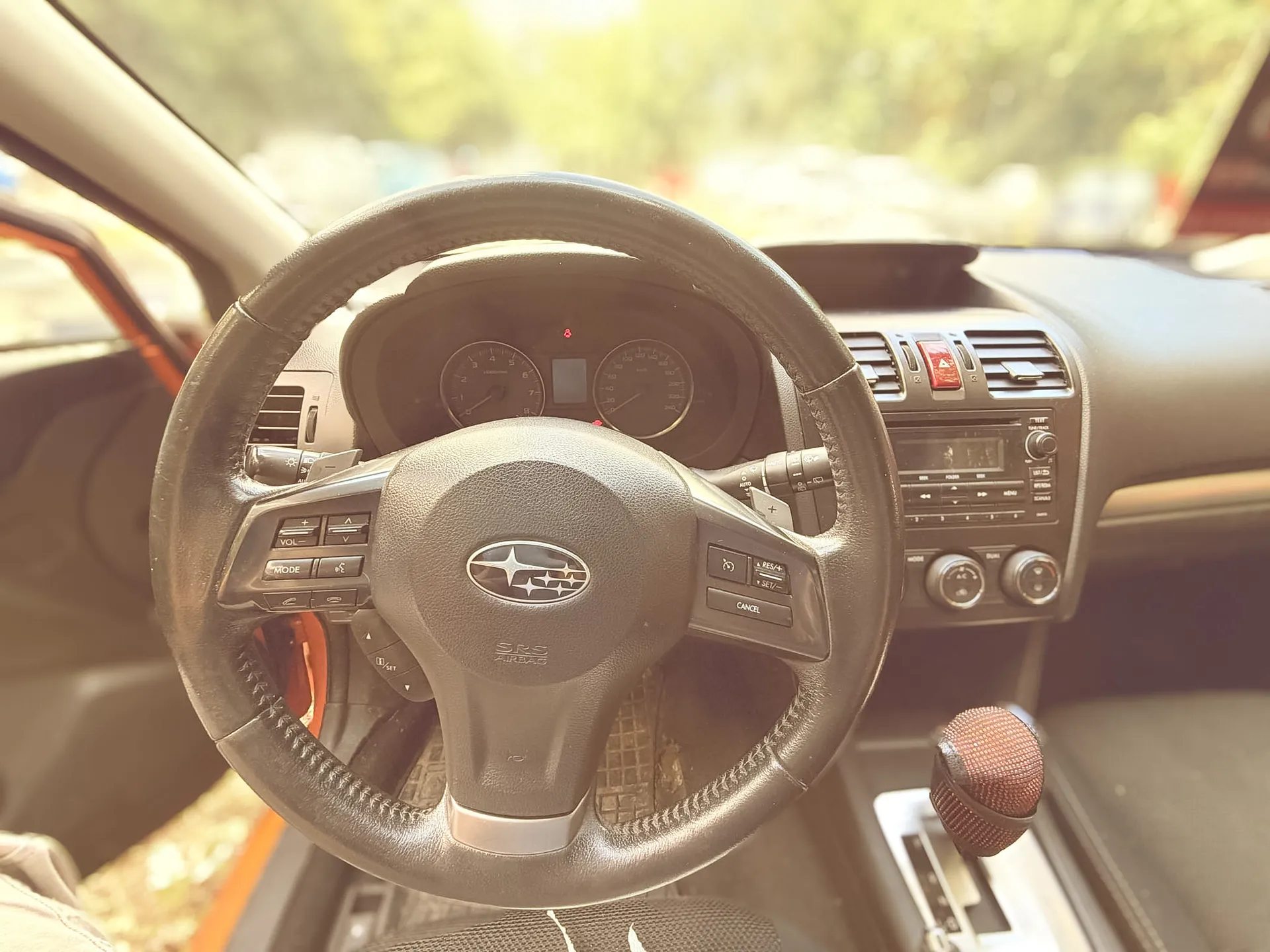 Wnętrze samochodu marki Subaru z perspektywy kierowcy. Widoczna jest skórzana kierownica z logo Subaru na środku i napisem 'SRS AIRBAG'. Za kierownicą znajduje się panel wskaźników z prędkościomierzem i obrotomierzem. Po prawej stronie widoczna jest konsola środkowa z nawiewami, przyciskiem alarmowym oraz radiem. W dolnej części obrazu widoczna jest lewarek zmiany biegów z czerwoną osłoną gałki.