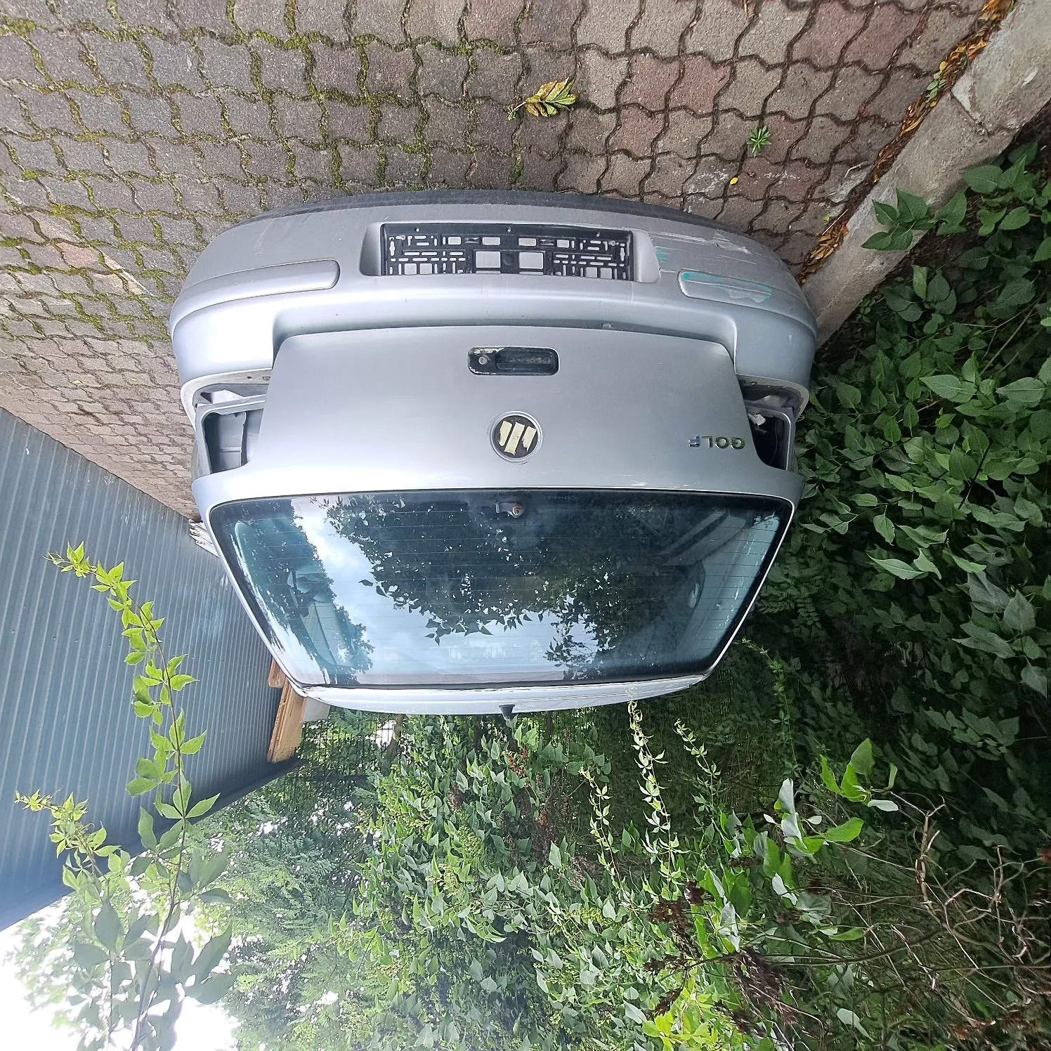 Srebrny przód samochodu marki Volkswagen Golf, który leży na boku na kostce brukowej. Widoczna jest przednia szyba, maska z logo marki oraz napis GOLF. W tle znajdują się zielone krzewy i fragment niebieskiego zadaszenia.