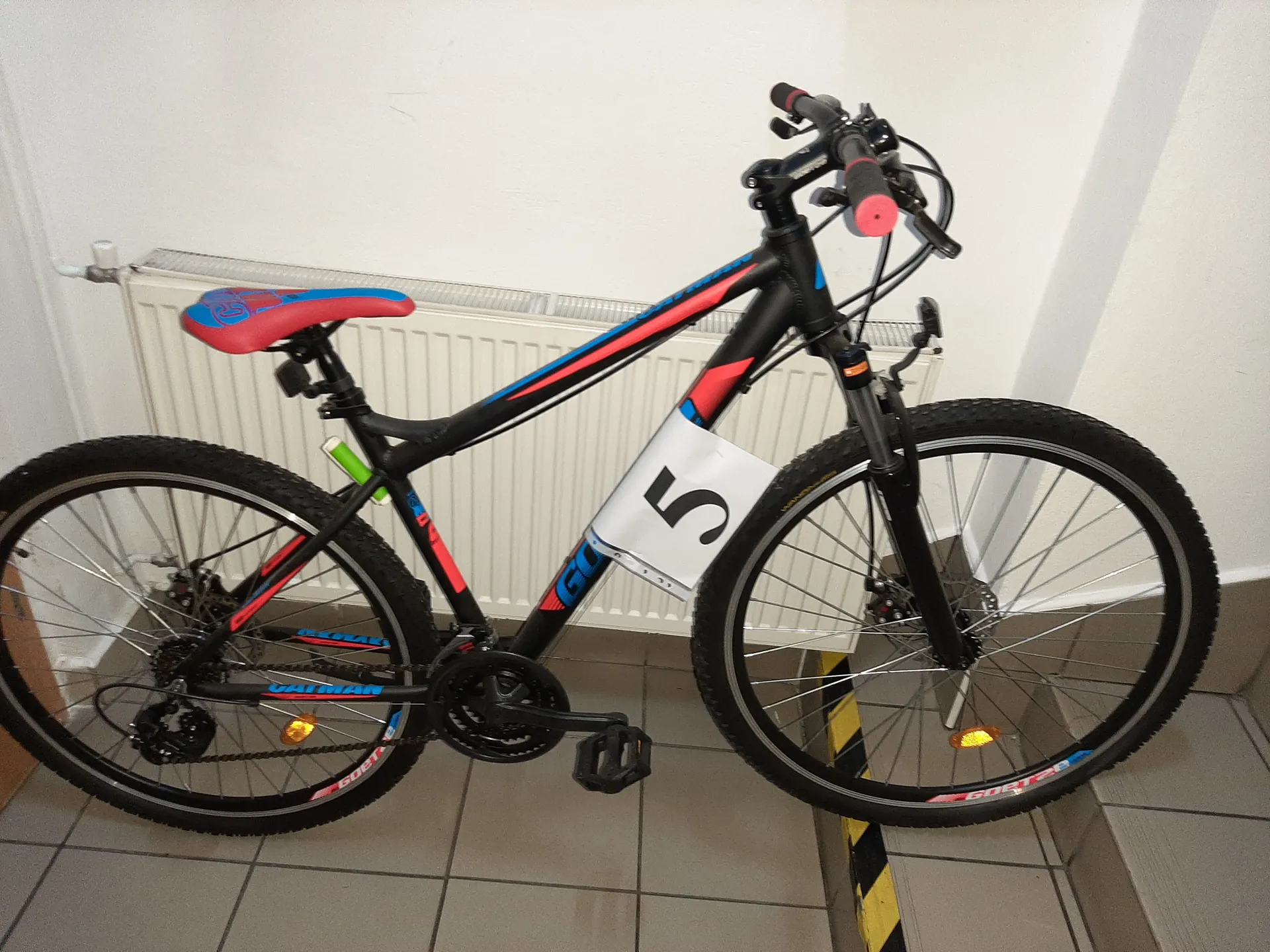 Czarny rower z czerwonymi i niebieskimi akcentami, z czerwonym siedziskiem i różowymi uchwytami na kierownicy. Na ramie roweru umieszczono białą tabliczkę z czarną cyfrą 5. Rower stoi na podłodze z szarymi płytkami, na tle białego grzejnika. Widoczne są pomarańczowe odblaski na kołach oraz zielony element na ramie.