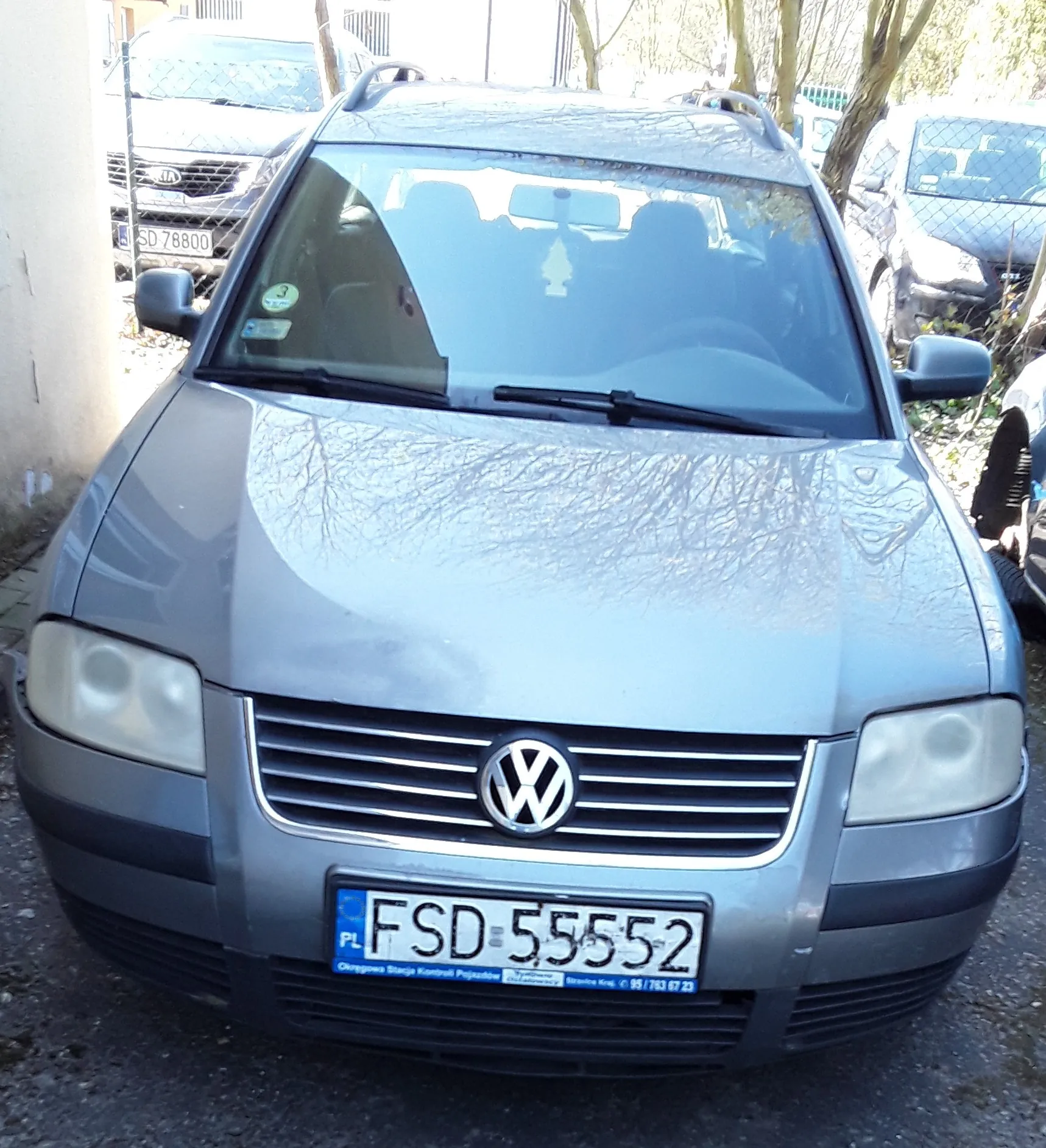Przedni widok szarego samochodu marki Volkswagen z tablicą rejestracyjną FSD 55552. Na tablicy widoczny jest niebieski pas z napisem PL oraz dodatkowy napis na dole. W tle znajdują się inne pojazdy, ogrodzenie siatkowe, drzewa bez liści i fragment budynku po lewej stronie. Na przedniej szybie widoczne są naklejki, a na masce odbicie gałęzi drzew.
