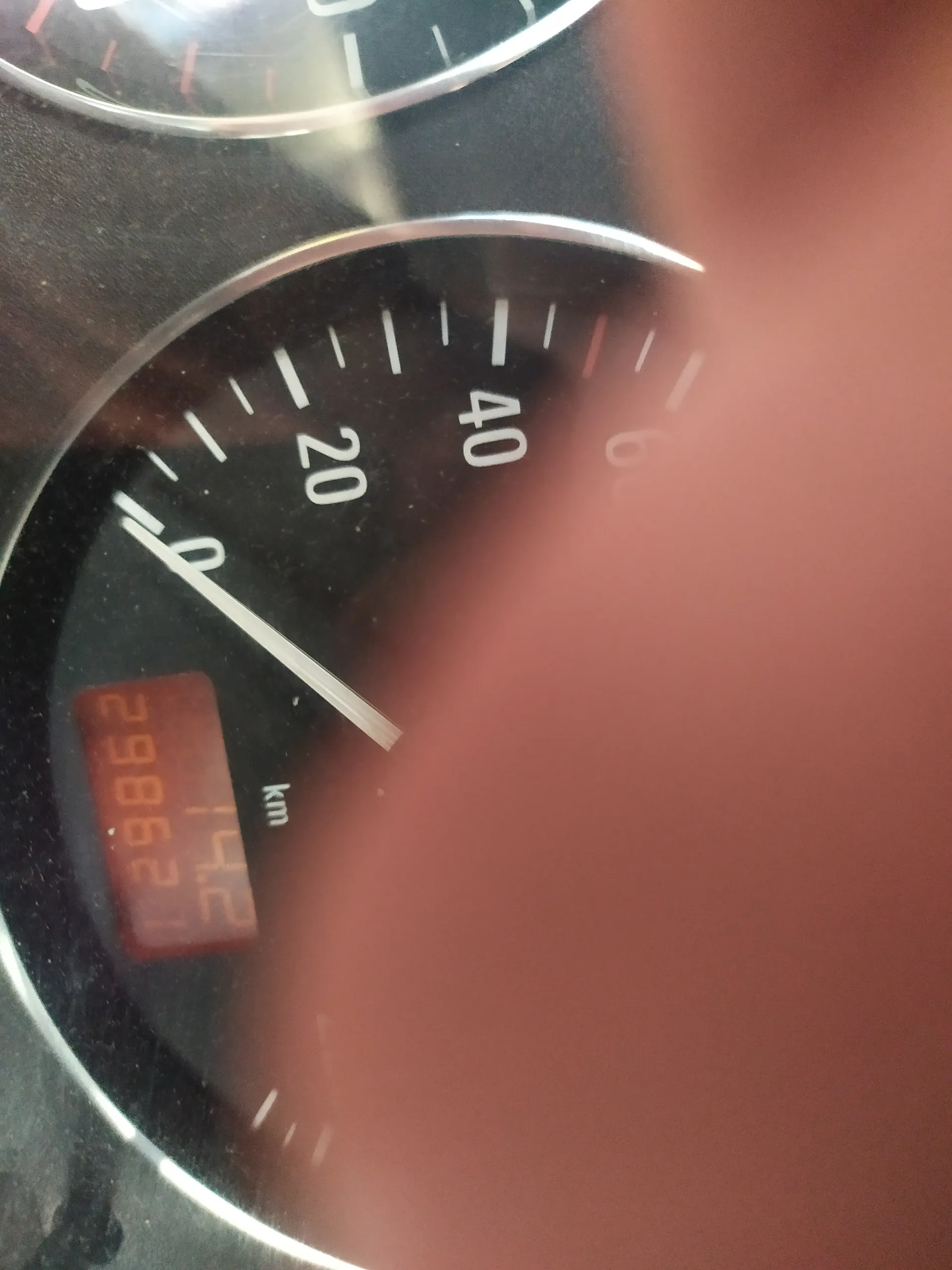 Zbliżenie na samochodowy wskaźnik prędkości z czarnym tłem i białymi znacznikami. Widoczne liczby 0, 20, 40, 60 oraz wskaźnik wskazujący blisko zera. Poniżej znajduje się czerwony wyświetlacz cyfrowy z napisem '14.2' i '29862.1', obok oznaczenie 'km'. Część obrazu przykrywa palec. Tło stanowi ciemna deska rozdzielcza.