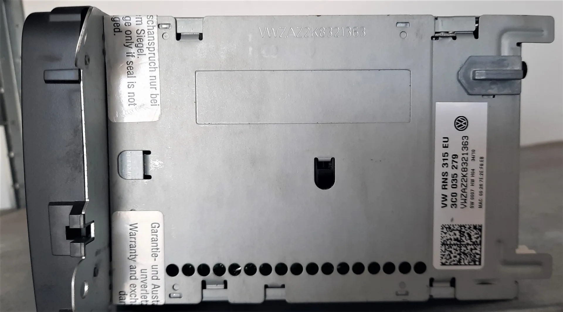 Srebrna obudowa urządzenia elektronicznego, prawdopodobnie radia samochodowego. Na obudowie znajdują się naklejki z tekstem. Jedna z nich zawiera napis: 'VW RNS 315 EU', '3C0 035 279', 'VWZA22K8321363', 'SW 0007 HW HIA 34/10', 'MAC: 00 20 7E 2E FB EB' oraz kod QR. Inna naklejka zawiera tekst w języku niemieckim i angielskim dotyczący gwarancji: 'Garantie- und Austausch', 'Warranty and exchange'. Na górnej krawędzi obudowy widnieje numer seryjny: 'VWZA22K8321363'.