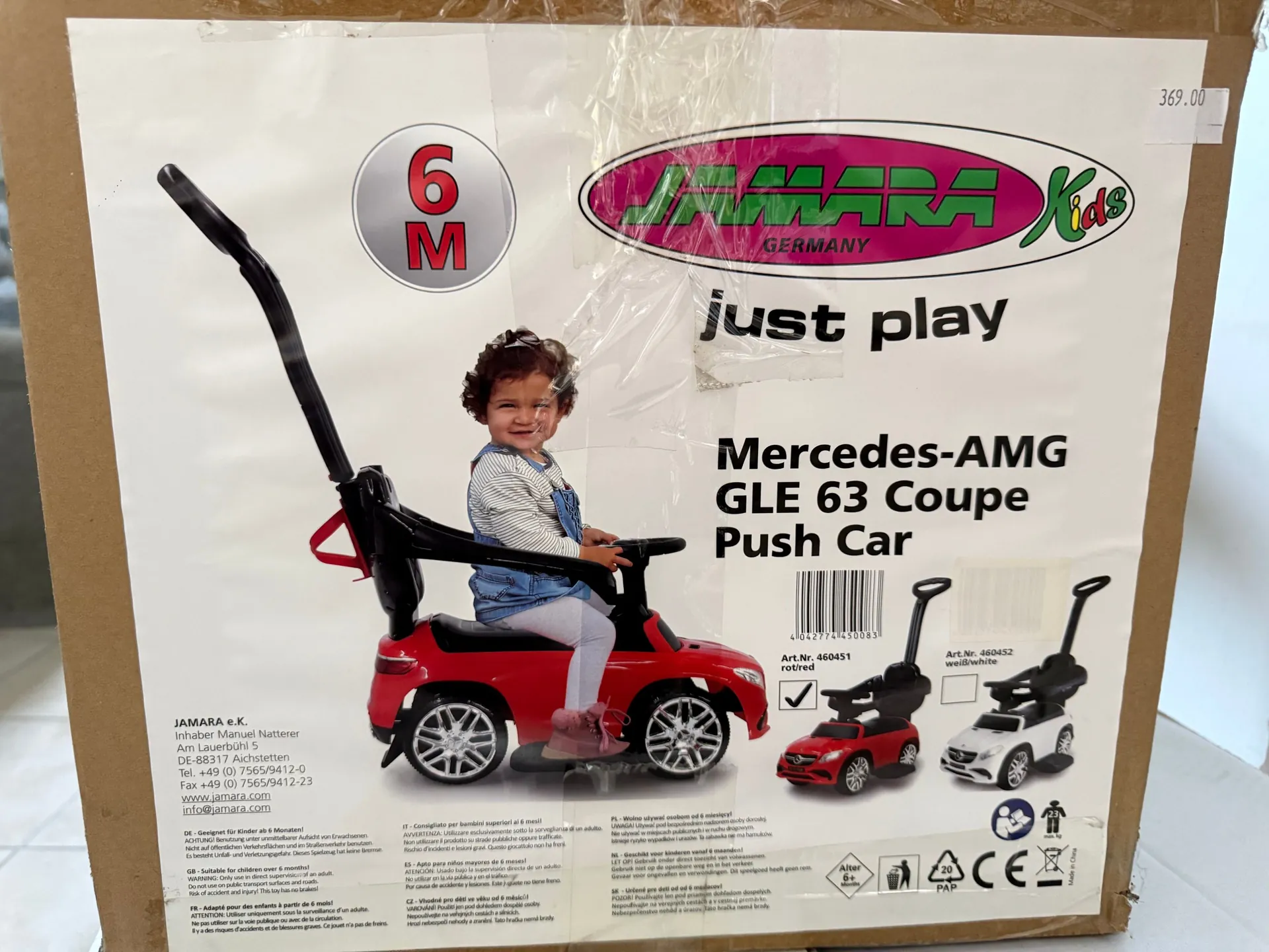 Kartonowe opakowanie zabawki Jamara Kids. Na przodzie znajduje się zdjęcie dziecka siedzącego w czerwonym samochodziku elektrycznym Mercedes-AMG GLE 63 Coupe Push Car, który posiada czarny uchwyt do pchania dla opiekuna. W górnej części widnieje logo 'JAMARA Kids GERMANY', napis 'just play' oraz ikona '6M' w kółku. Poniżej znajduje się nazwa modelu samochodu oraz kody kreskowe i numery katalogowe. W dolnej części opakowania widnieją instrukcje i ostrzeżenia w wielu językach oraz dane kontaktowe firmy Jamara e.K. W prawym górnym rogu naklejona jest biała etykieta z ceną '369.00'.