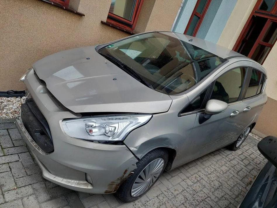 Srebrny samochód osobowy z widocznymi uszkodzeniami przodu. Maską jest podniesiona i wygięta, przedni zderzak jest pęknięty i częściowo oderwany, a przednia szyba jest rozbita. Auto stoi na kostce brukowej przed budynkiem z beżową elewacją i oknami w brązowych ramach.