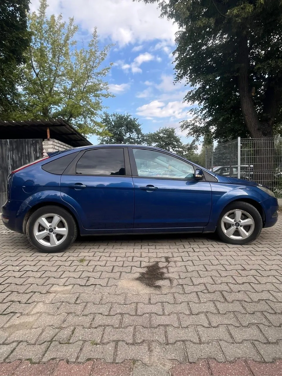 Niebieski samochód osobowy marki Ford Focus zaparkowany bokiem na kostce brukowej. W tle widoczne są zielone drzewa, fragment drewnianego ogrodzenia oraz niebo z białymi chmurami.