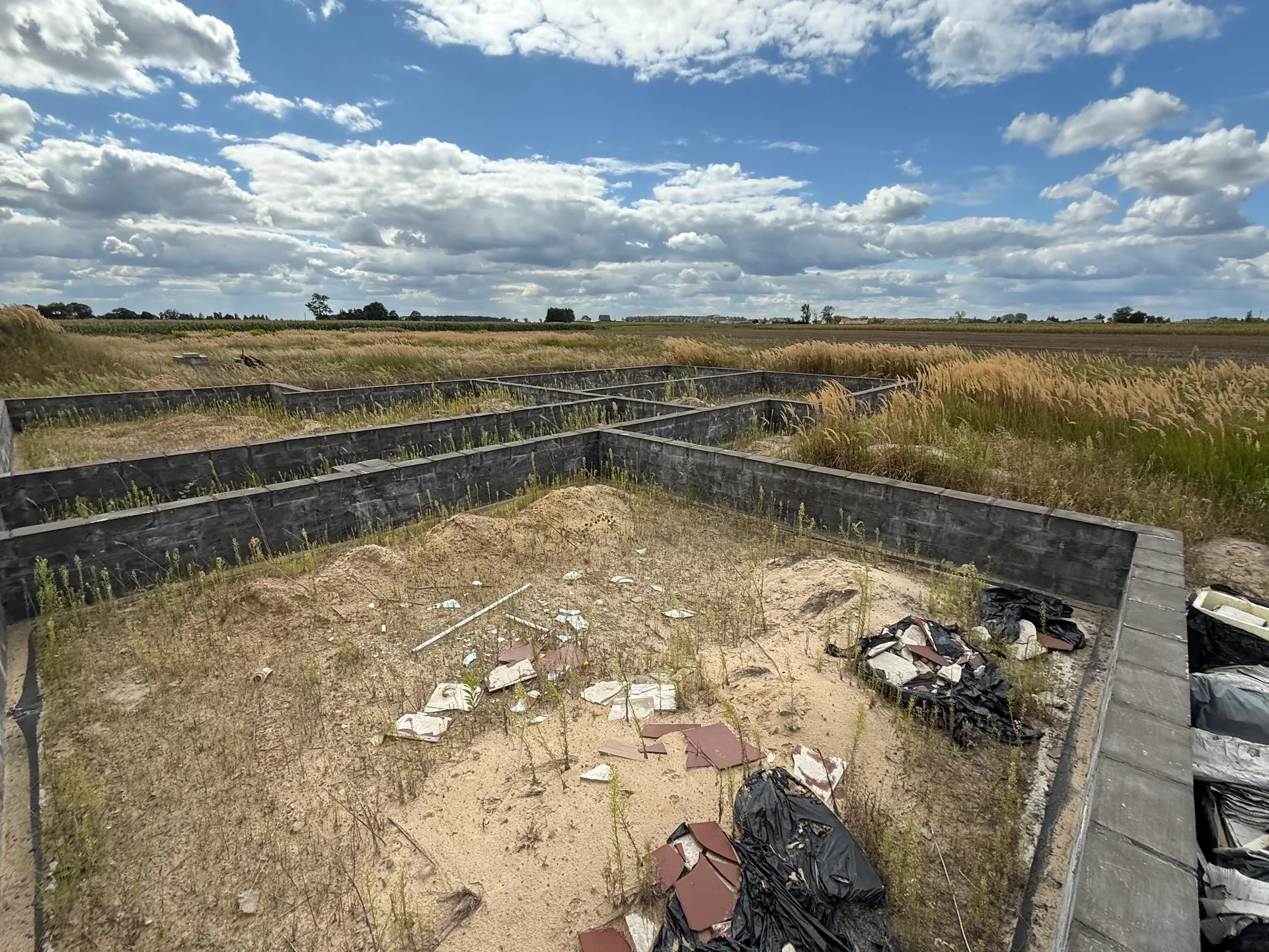 Widok z góry na fundamenty budynku w formie betonowych ścian oporowych, tworzących prostokątne pomieszczenia. Wnętrze fundamentów wypełnione jest piaskiem, gruzem budowlanym oraz czarną folią budowlaną. W tle rozciąga się płaski krajobraz z wysokimi trawami i polami pod niebem z rozproszonymi białymi chmurami.