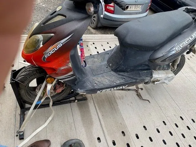 Zdjęcie przedstawia czerwono-czarny skuter marki FOCUS zaparkowany na metalowej platformie z otworami. Na przedniej części skutera widoczny jest napis 'FOCUS', a na podłodze 'KIPWAY'. W tle znajduje się szary samochód z tablicą rejestracyjną 'W1 80HG'. Na dole zdjęcia widoczna jest część stopy osoby w brązowym obuwiu. Skuter jest przymocowany białą taśmą do platformy.