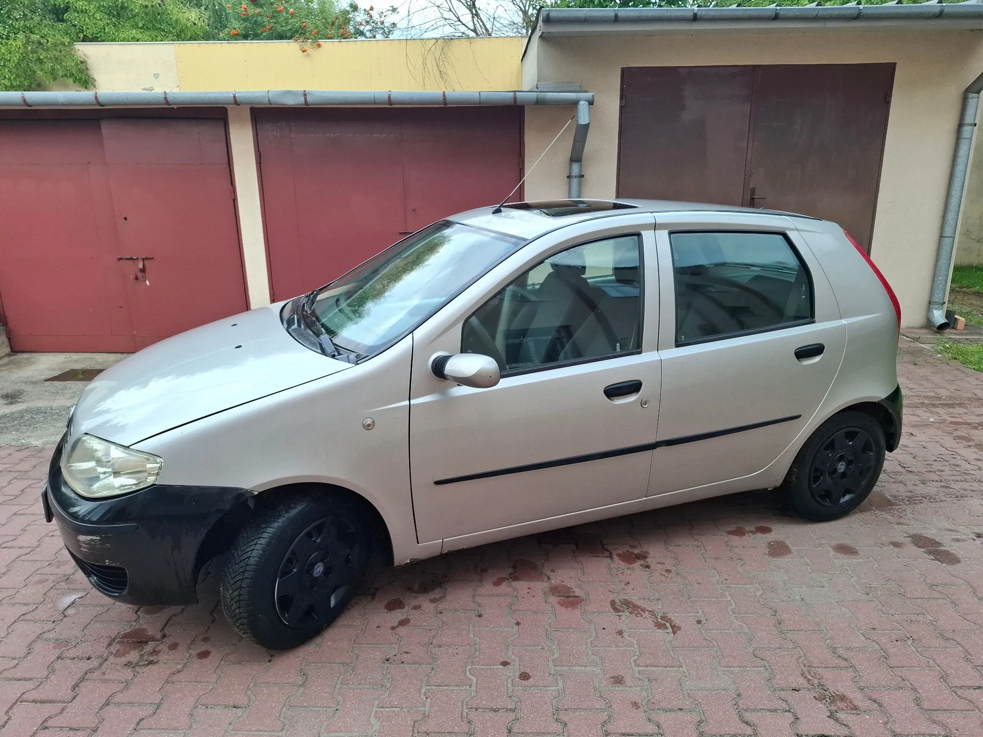 Srebrny, pięciodrzwiowy samochód osobowy zaparkowany na kostce brukowej przed garażami z brązowymi drzwiami. Auto ma czarne felgi i czarny przedni zderzak.