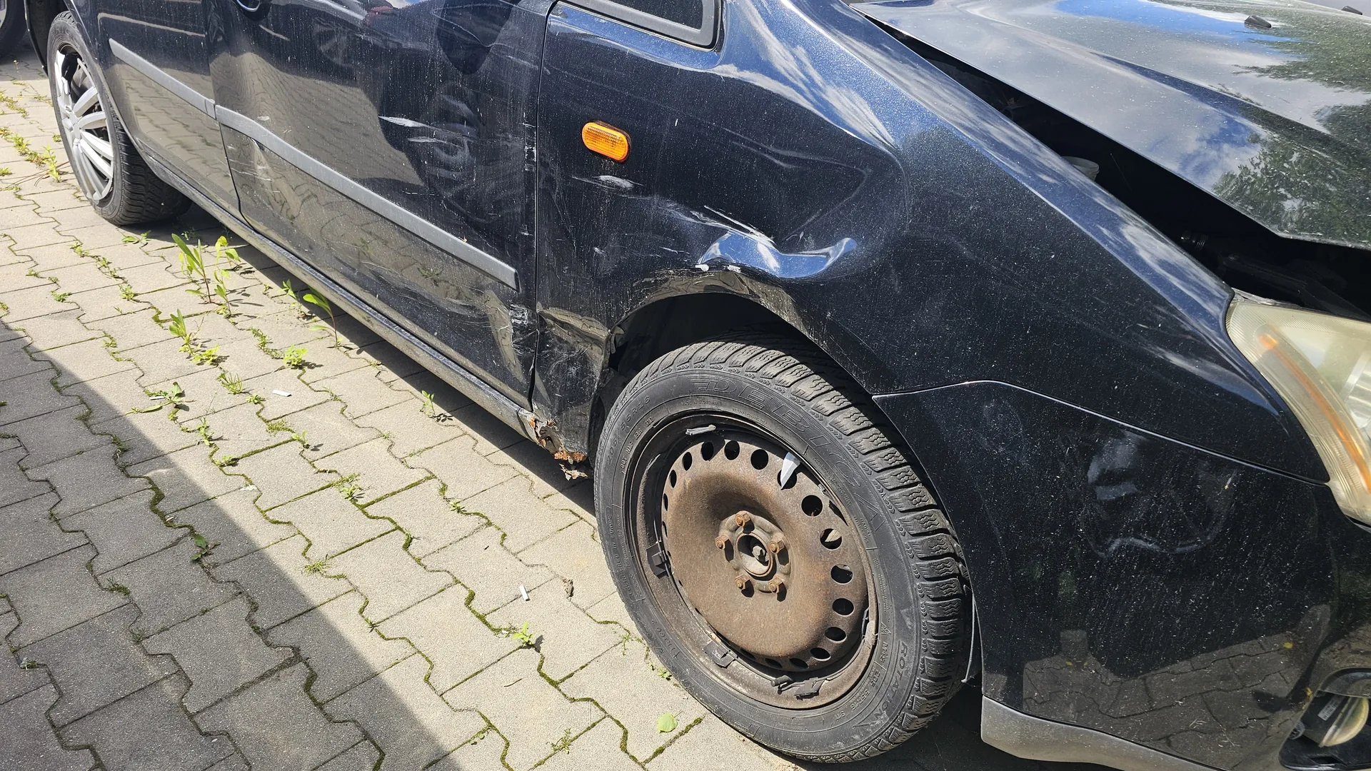 Zdjęcie przedstawia czarny samochód osobowy z widocznymi uszkodzeniami na przedniej prawej stronie, w tym wgnieceniami nad kołem i pod maską. Przednie prawe koło ma zardzewiałą felgę i zużyty oponę. Samochód stoi na chodniku z betonowych płyt, między którymi rosną zielone rośliny. W tle widoczne są odbicia drzew i nieba na błyszczącej karoserii.
