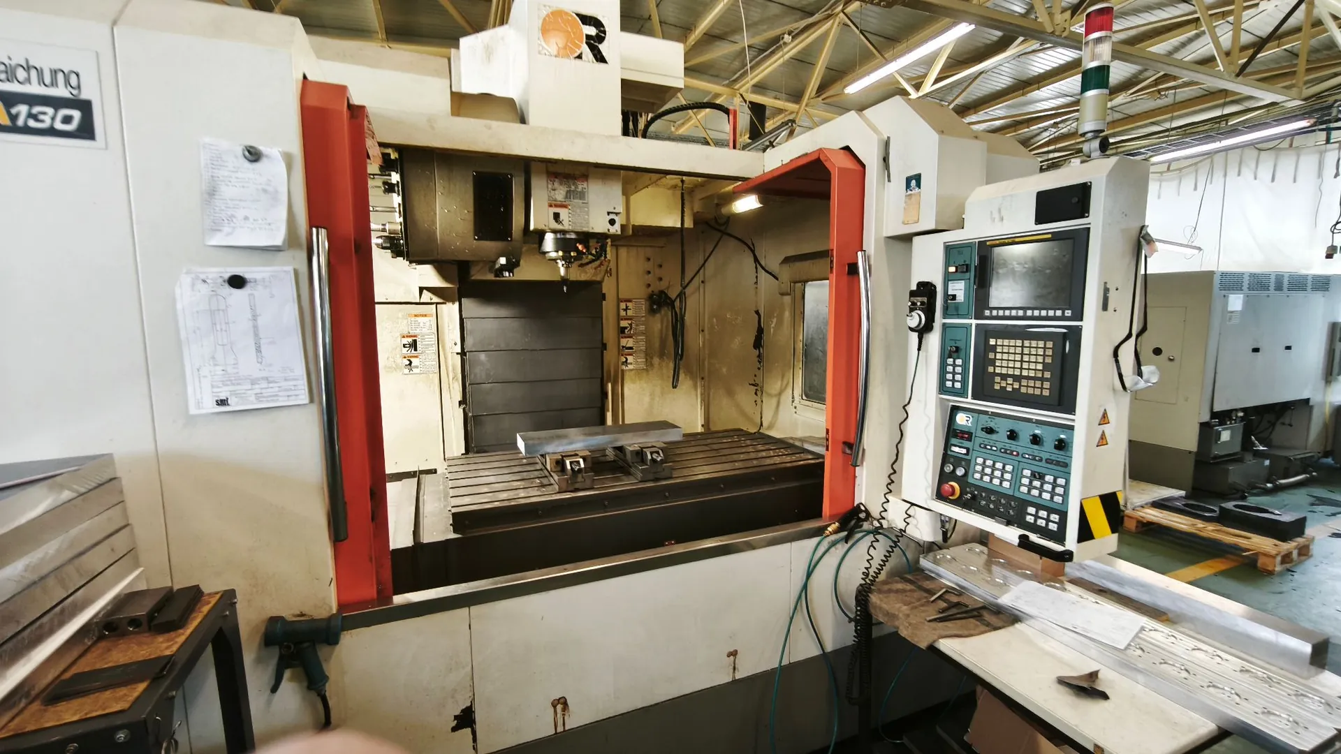 Duża, biała maszyna CNC do obróbki metalu w hali przemysłowej. Maszyna posiada otwartą komorę roboczą z metalowym stołem, panel sterowania z ekranem i przyciskami po prawej stronie oraz czerwone obramowanie otworu. Po lewej stronie obudowy przyklejone są kartki z dokumentacją techniczną. W tle widoczna jest konstrukcja dachu hali i inne urządzenia warsztatowe.