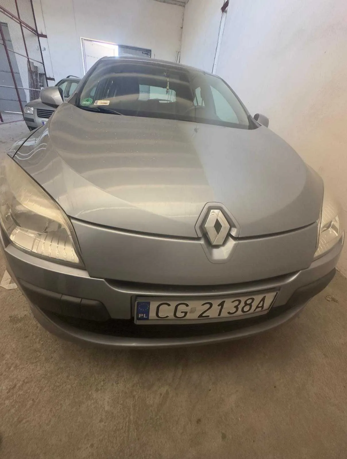 Przedni widok srebrnego samochodu marki Renault z tablicą rejestracyjną CG 2138A z oznaczeniem PL. W tle widoczny inny samochód, białe ściany i betonowa podłoga w pomieszczeniu. Na przedniej szybie naklejki, a na masce logo Renault.