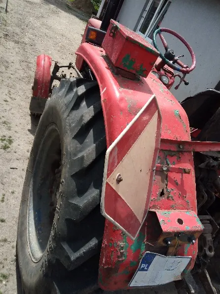 Zdjęcie przedstawiające tylną część czerwonego traktora z widocznym dużym, czarnym kołem z bieżnikiem. Na tylnym błotniku zamocowany jest biało-czerwony znak ostrzegawczy w kształcie rombu. Na dole znajduje się biała tabliczka z napisem 'PL'. Traktor jest w widocznym stanie zużycia z odpryskami farby.
