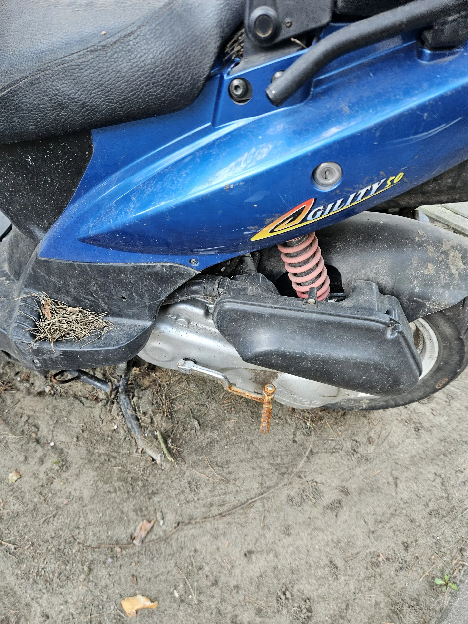 Zbliżenie na niebieski skuter z widocznym logo "Galaxy 50" w kolorach żółtym, czerwonym i białym. Widoczne są czarne elementy nadwozia, różowa sprężyna, czarna opona oraz metalowe części. Na tle brudnej ziemi z suchymi liśćmi i gałązkami. Skuter pokryty jest kurzem i śladami brudu.