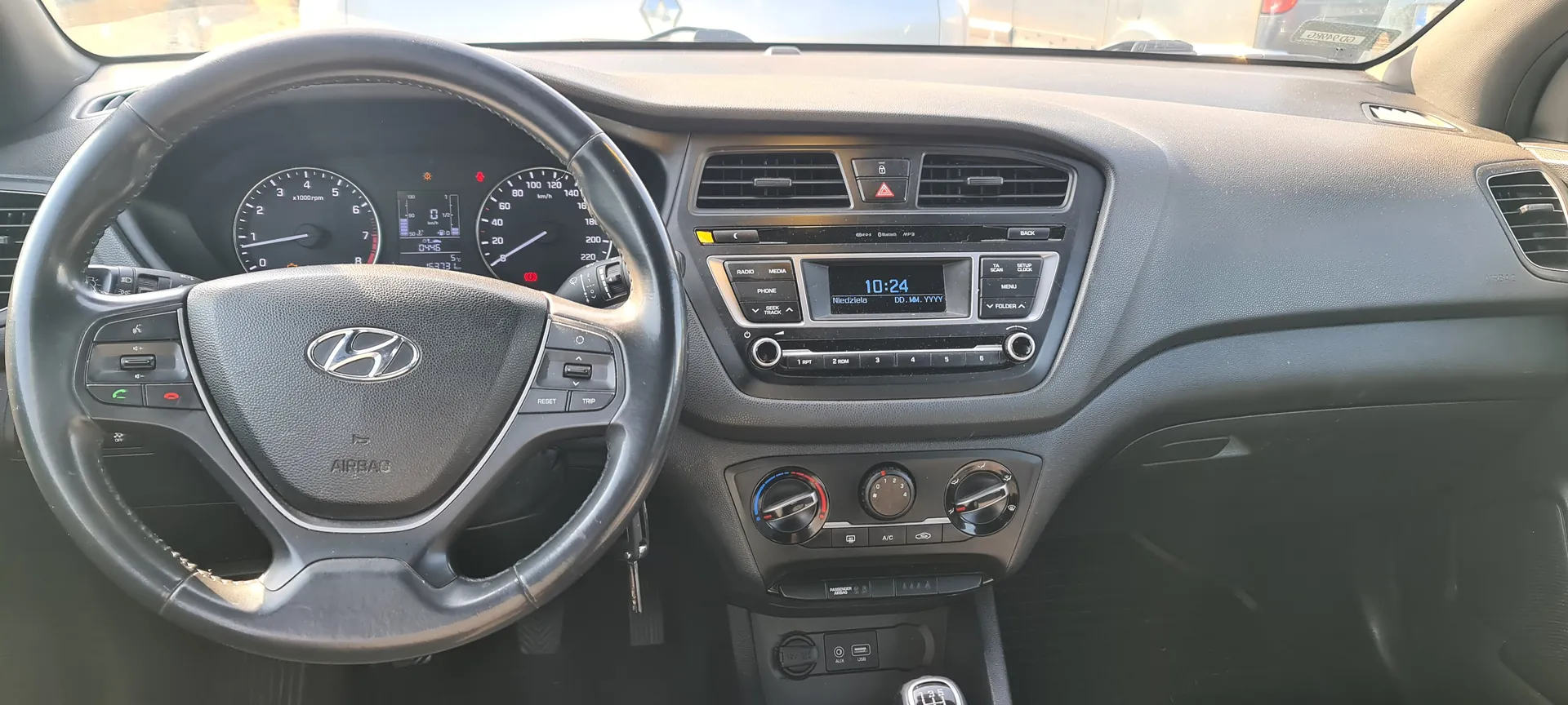 Wnętrze samochodu marki Hyundai z widoczną kierownicą z logo Hyundai, licznikami prędkości i obrotomierzem, centralnym wyświetlaczem pokazującym godzinę 10:24 oraz dzień tygodnia Niedziela. Deska rozdzielcza w kolorze szarym z przyciskami sterującymi klimatyzacją i systemem audio. Widoczne są również pokrętła regulacji klimatyzacji oraz dźwignia zmiany biegów.