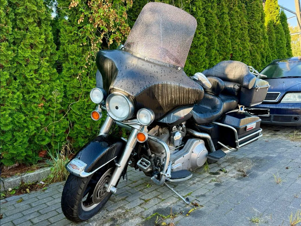 Czarny motocykl typu touring z dużą owiewką i wysoką szybą ochronną, zaparkowany na kostce brukowej przed zielonym żywopłotem z tuj. Motocykl posiada dwa przednie reflektory, chromowane elementy silnika i wydechu oraz dwa boczne kufry. W tle po prawej stronie widoczny jest fragment niebieskiego samochodu.
