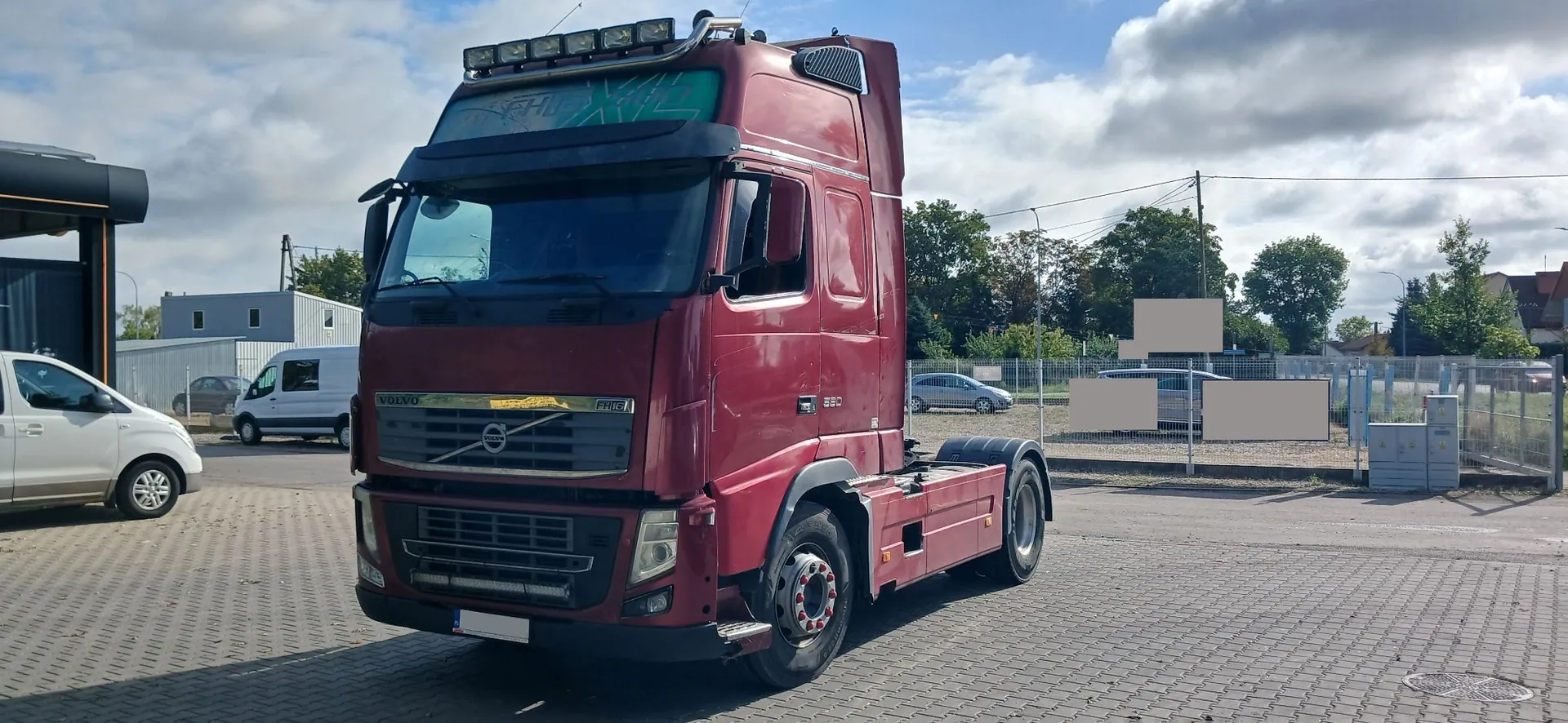 Czerwony ciągnik siodłowy marki Volvo zaparkowany na brukowanym placu. Pojazd posiada dodatkowe oświetlenie na dachu oraz ciemne szyby. W tle widoczne są białe samochody dostawcze, szary budynek, ogrodzenie z siatki oraz drzewa pod zachmurzonym niebem.