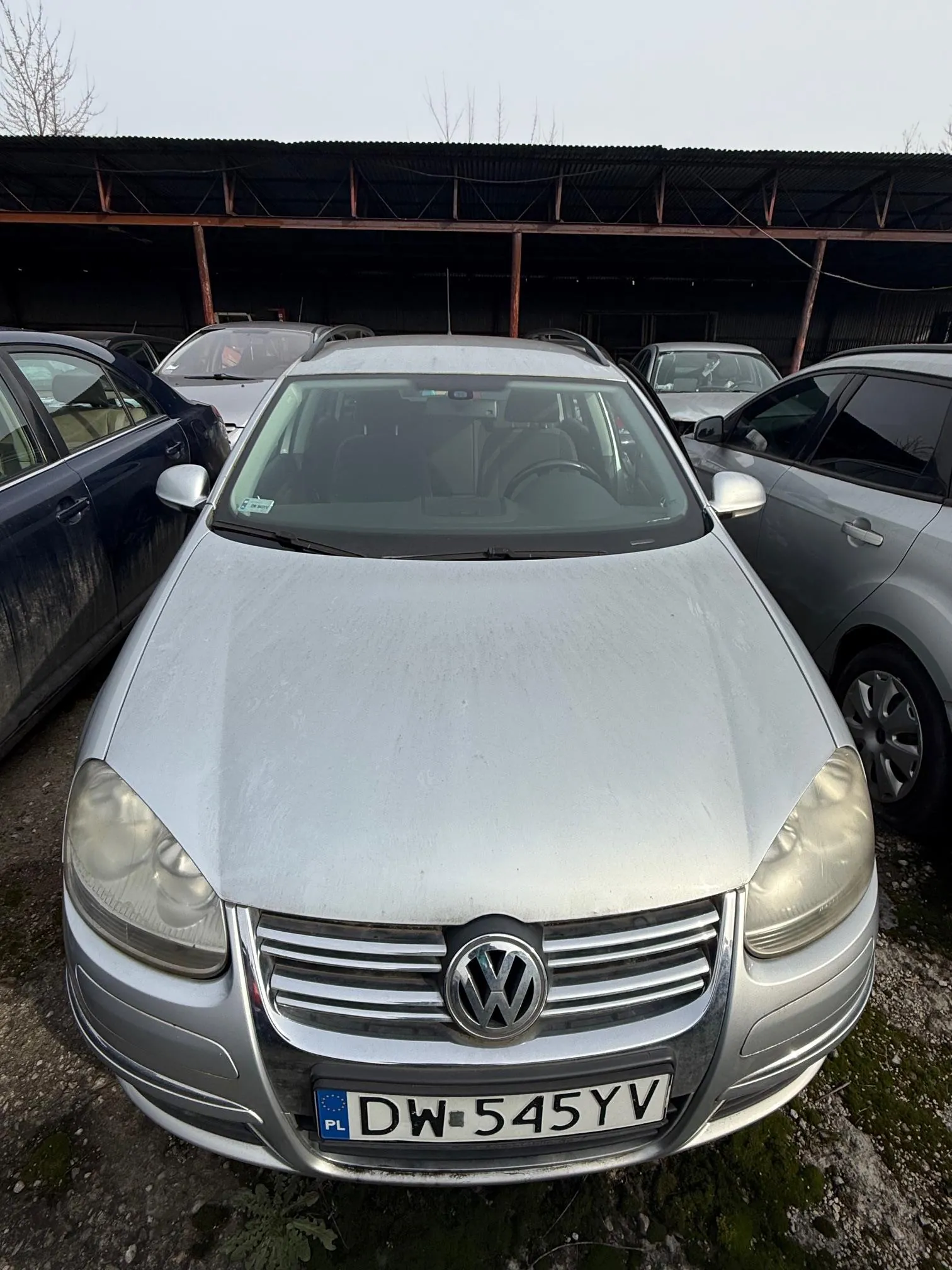 Srebrny samochód marki Volkswagen z tablicą rejestracyjną DW 545YV (PL) na pierwszym planie. W tle widoczne są inne pojazdy, metalowa konstrukcja dachu oraz ziemia z drobnoziarnistym podłożem i mchem. Samochód stoi na otwartym placu, a na szybie przedniej znajduje się mała naklejka.