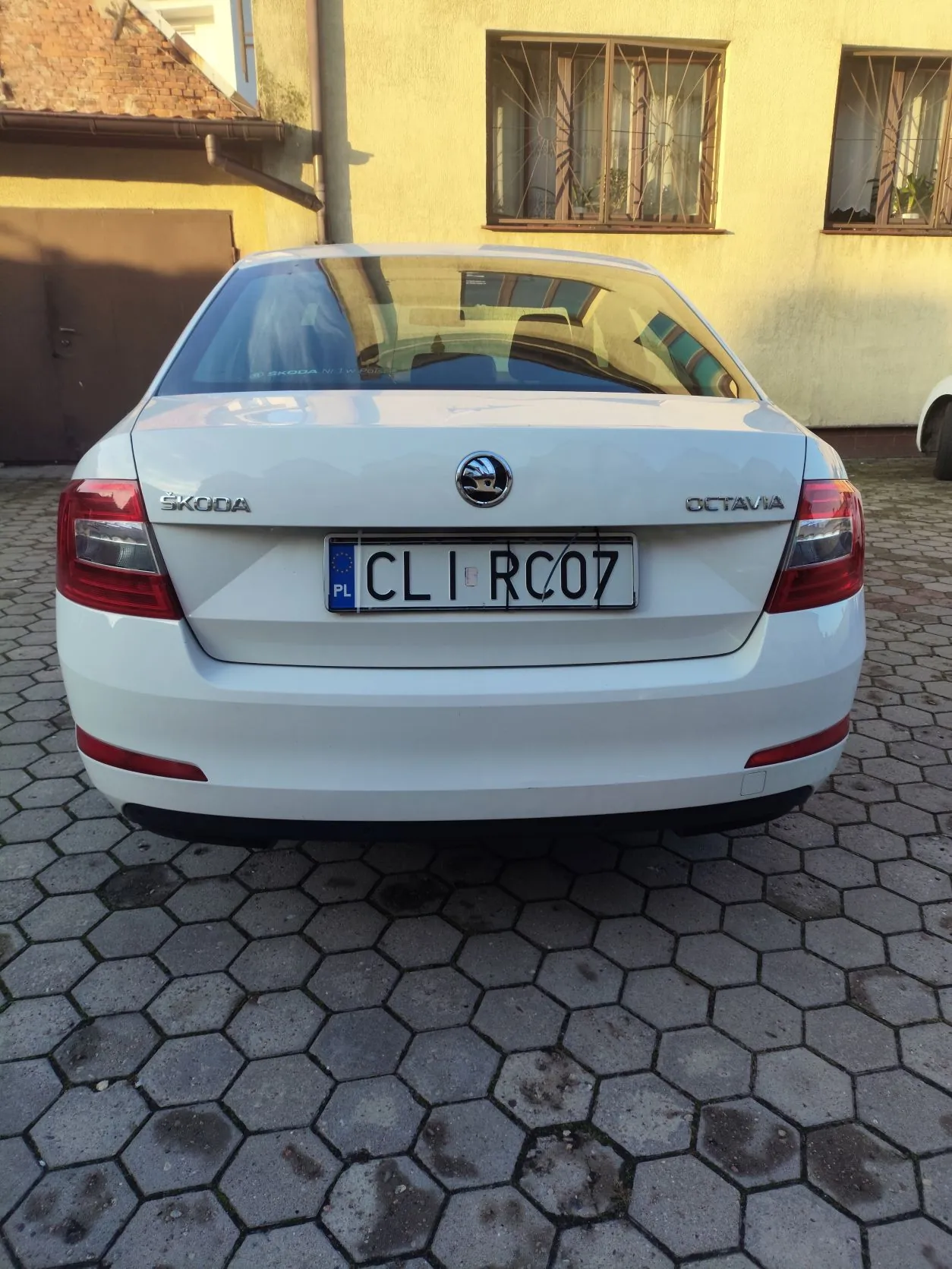 Tylny widok białego samochodu marki Skoda Octavia zaparkowanego na kostce brukowej o sześciokątnym kształcie. Samochód ma polską tablicę rejestracyjną o numerze CLI RC07. W tle widoczny jest fragment żółtego budynku z dwoma oknami zabezpieczonymi kratami oraz brązowa brama garażowa.